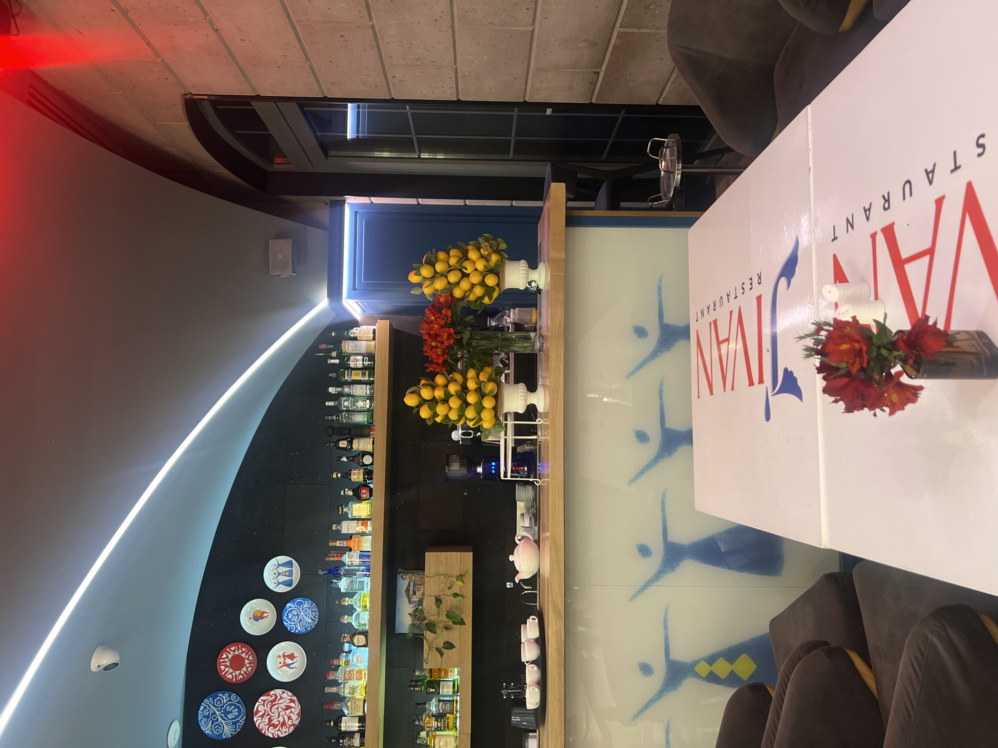This is an interior view of the Jivan Restaurant in Yerevan, Armenia. The restaurant features a bar area with a curved black wall displaying shelves stocked with bottles, likely alcoholic beverages. Decorative plates are mounted on the wall below the shelves. In front of the bar is a counter or serving area adorned with flowers and fruit displays. A white table prominently features the restaurant's name, "Jivan Restaurant," in red and blue lettering. A vase of red flowers sits on the table. Brown upholstered chairs are visible around the table. The overall design appears modern and stylish. The wall behind the bar is decorated with blue stylized figures.