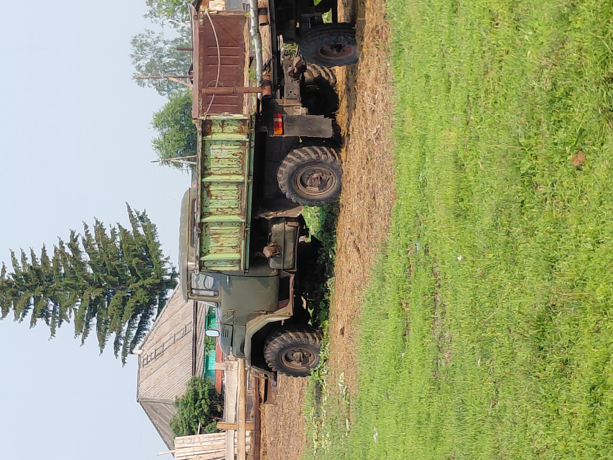 An exterior, full shot captures an olive green, vintage truck with a trailer parked on a dirt and grass area. The truck is positioned facing right, with its front slightly obscured by shadows. The truck features large, rugged tires and a weathered, green cargo bed. The trailer behind it is also a rusty brown color and appears to be carrying some unseen cargo.
In the background, a large, dark green evergreen tree stands prominently. To the left of the tree, a house with a grey, corrugated roof is visible, with some wooden structures and greenery surrounding it. The sky above is a pale, hazy blue. The overall scene suggests a rural or semi-rural setting, likely in the city of Novosyolovo, Russia, given the context. There are no people present in the image, and the environment appears to be outdoors on a clear day.