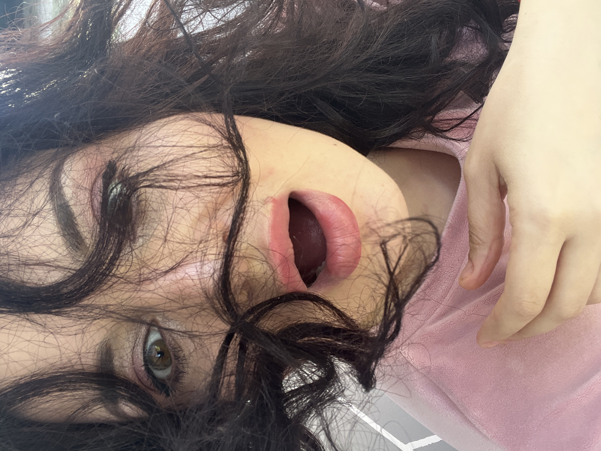 A close-up, high-angle shot captures a person, likely female, with a strong tilt to the perspective, giving the impression of looking down on them as they lie down. The dominant features are their dark, wavy hair, part of their face, and a hand resting on pink fabric.
The person has thick, dark brown or black hair that appears somewhat disheveled and frames their face, spreading across much of the image. One eye, visible on the left side, is open and appears to be a light brown or hazel color, with visible eyelashes and traces of eyeliner or eyeshadow. Their mouth is open, lips parted, revealing the inside of their mouth and tongue, with the lips themselves being a soft pinkish-red hue. Their skin tone is fair.
A hand, likely belonging to the same person, is visible on the right side of the frame. It has light skin and slightly curled fingers, resting on a soft, light pink fabric. This fabric appears to be part of clothing, possibly a top, or a blanket, with a plush texture like velour or fleece.
The lighting is bright, casting some highlights on the hair and skin. The background is mostly obscured by the hair and fabric, offering no specific location cues. There is no visible text. The person's expression is ambiguous due to the angle and open mouth, suggesting anything from a gasp to a sigh, or simply being relaxed.