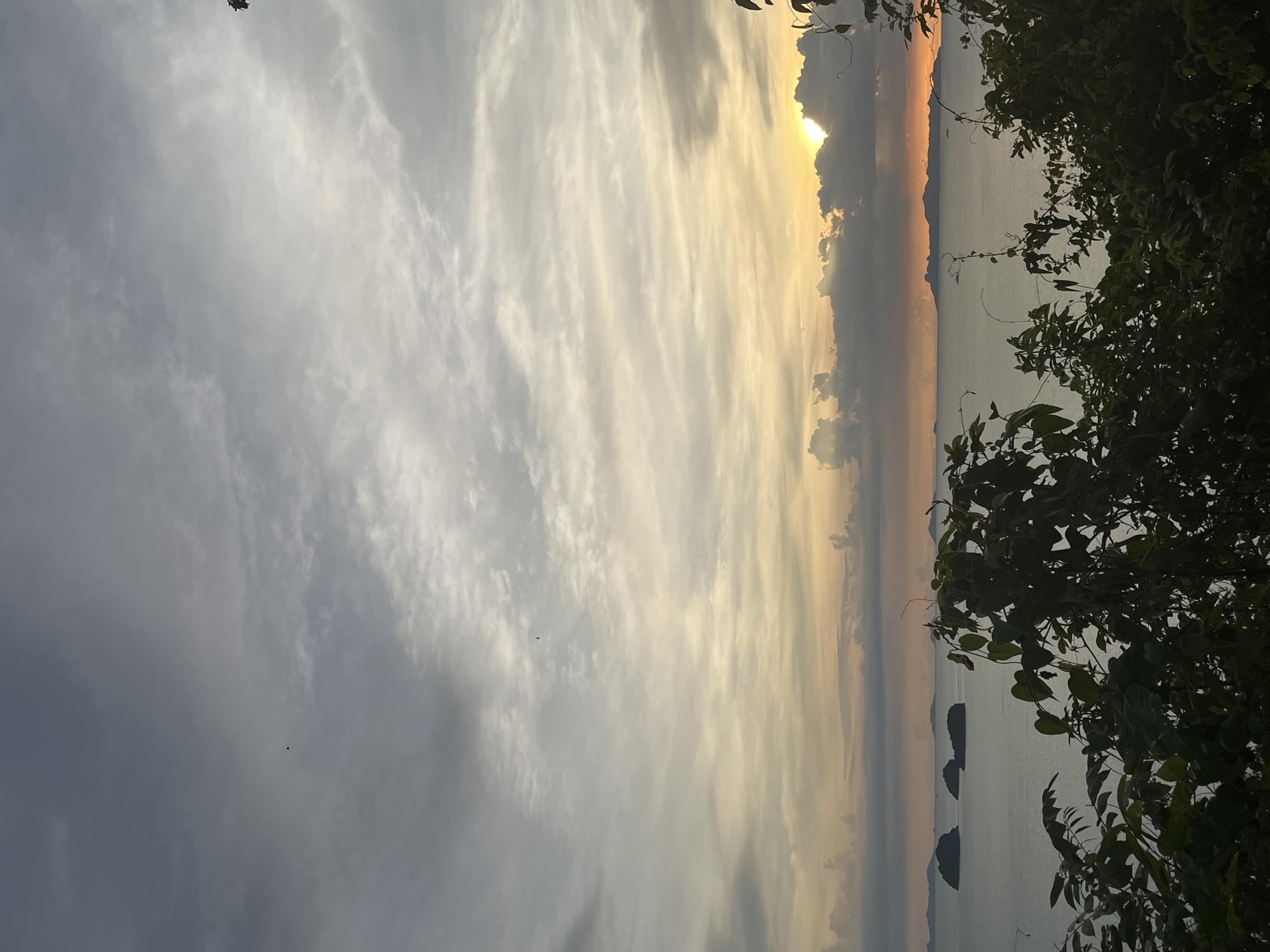 A sunset view from Baan Pang Ka, Thailand, shows islands in the distance across a calm sea. The sky is filled with dramatic clouds, with the sun partially obscured behind a dark cloud formation, casting a warm glow on the horizon. In the foreground, lush green foliage frames the scene, with branches and leaves reaching into the frame. The water is a muted blue-gray, reflecting the overcast sky. Several dark, rounded islands are visible in the middle ground, with a longer, more distant island range stretching across the horizon. The overall atmosphere is serene and picturesque.