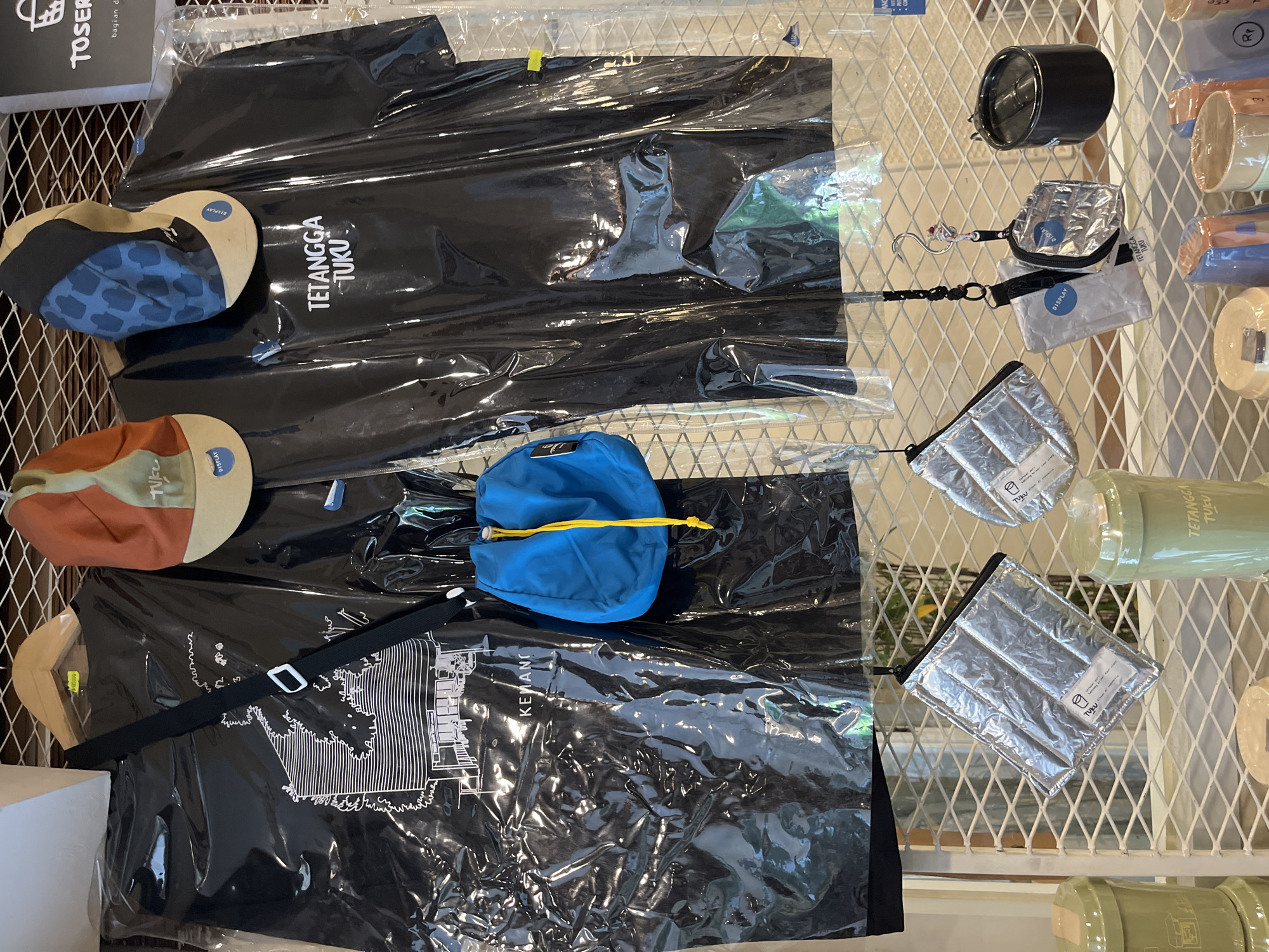This is a medium shot, eye-level, indoor photograph of merchandise displayed on a white wire rack against a tiled wall. The scene is likely in a store in West Jakarta, Indonesia, as indicated by the merchandise's branding. The primary subjects are two black t-shirts, each encased in a clear plastic sleeve. The t-shirt on the left features a white line drawing of a building and trees. The t-shirt on the right has white text that reads "TETANGGA TUKU". Both t-shirts are hung on wooden hangers.
In front of the t-shirts, a small, bright blue drawstring bag with a yellow cord is suspended. Above the t-shirts, two baseball caps are visible. The cap on the left is orange and tan with a blue pattern on the front. The cap on the right is blue with a tan brim and a similar blue pattern. Both caps have small circular "DISPLAY" tags.
Below the t-shirts, several smaller items are displayed on the wire rack. There are at least three quilted, silver pouches with zippers. One of these pouches has a label with text and a small drawing of a cup. Two of these pouches are suspended by black cords, one of which has a small keyring attached. There is also a white pouch with a blue "DISPLAY" tag. To the right, a black cylindrical container is visible, and next to it, a silver metallic pouch with a black zipper.
The background shows a textured tiled wall. In the lower portion of the image, part of another shelf is visible, displaying various cylindrical containers. Some of these containers are light green and have labels, including one with the text "TETANGGA TUKU". Other containers appear to be in various shades of orange, blue, and beige, some wrapped in plastic. The overall impression is of a retail display of clothing and accessories.