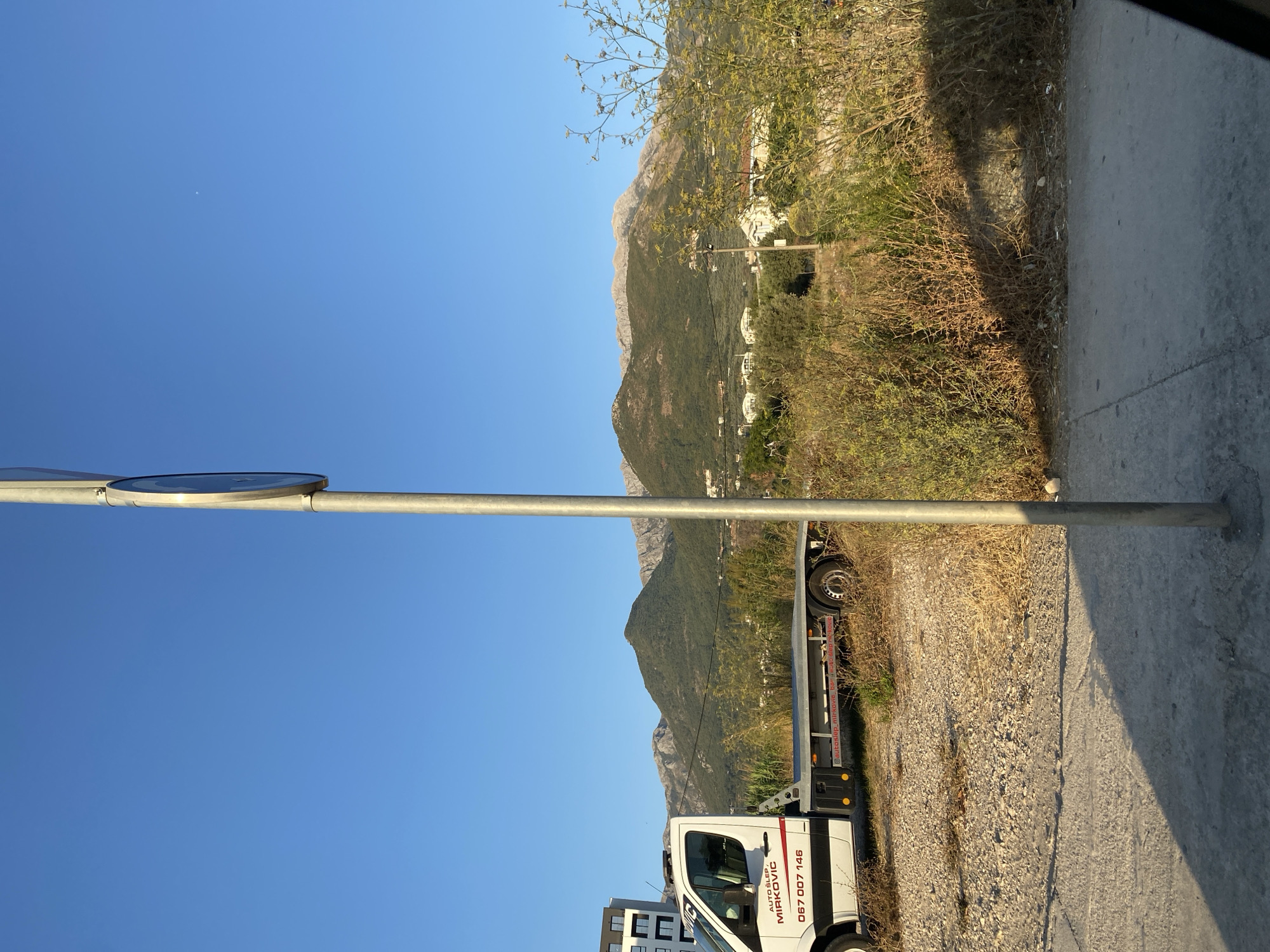 This image, captured on a clear day, depicts a tow truck parked on a gravelly surface in Bar, Montenegro. The truck, with "AUTO SLEP MIRKOVIC" and a phone number visible on its side, is positioned in front of a backdrop of lush green hills and mountains, some of which are partially obscured by the bright sunlight. A modern building can be seen to the left of the truck. A prominent metal pole, likely supporting a street sign, stands in the foreground, casting a shadow onto a concrete pathway. The environment appears somewhat unkempt, with dry vegetation and scattered debris visible around the truck and pathway. The sky is a vibrant, cloudless blue.