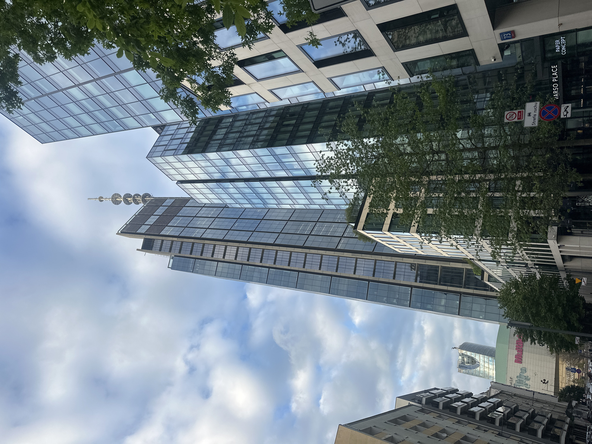 This is a ground-level, low-angle exterior shot of a street in Warsaw, Poland, focusing on modern architecture. The sky is overcast with scattered white clouds. Prominent in the foreground is a light-colored building with rectangular windows and dark frames. To its right is a tall, slender skyscraper clad in glass, reflecting the sky and surrounding buildings. To the left of this skyscraper, another glass-clad building is partially visible, and further left, a darker, older building with balconies lines the street. In the background, a distinctive curved skyscraper with a green roof is visible, as well as another tall tower with a lattice antenna. Several trees with green foliage are scattered throughout the scene, with one particularly large tree in the mid-ground obscuring parts of the buildings. Various street signs are visible, including traffic regulations and building names like "VARSO PLACE" and "PAPER CONCEPT". Cars and possibly a tram are visible on the street in the lower left portion of the image. The overall impression is one of a dense urban environment with a mix of architectural styles, dominated by the imposing modern skyscrapers.