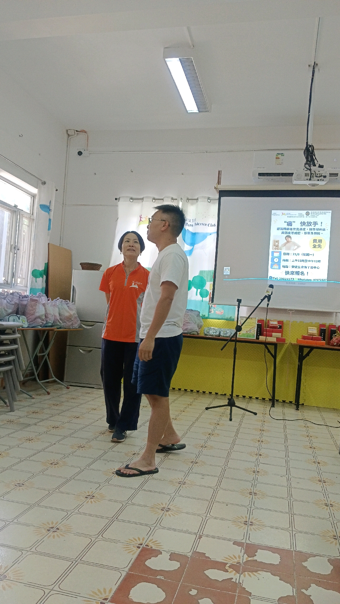 This image shows two people standing in what appears to be a community hall or classroom. On the left, a woman in an orange shirt and dark pants stands facing a projector screen. She has short dark hair and is looking upwards. To her right, a man in a white t-shirt and dark shorts is also looking upwards. He has short dark hair and glasses.
The room has tiled flooring with a floral pattern. Against the wall on the left are tables laden with bagged items. A refrigerator is also visible behind the woman. The projector screen displays text and images, including the words "痛" and "快放手!". There is a microphone on a stand in front of the screen. The overall lighting suggests an indoor setting during the day.
Based on the presence of the text "快放手!" (meaning "Let go quickly!") and "費用全免" (meaning "Free of charge"), as well as Chinese characters and phone numbers, this scene is likely located in Hong Kong, China, where Cantonese is spoken and such community outreach programs are common.