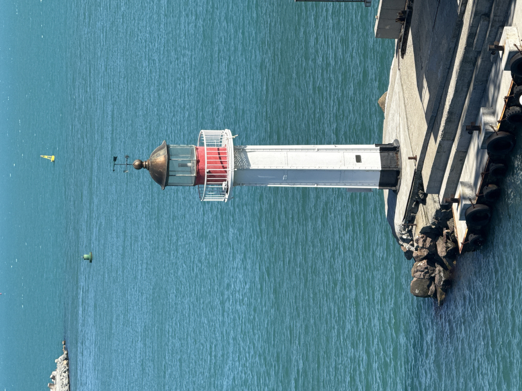 The image shows a lighthouse on a pier in Burgas, Bulgaria. The lighthouse is white with a red band around its lantern room, which is topped with a copper dome and a weather vane. The pier is made of concrete and is surrounded by rocks and the blue sea. In the background, there are buoys and a small yellow sailboat. The sky is clear and blue.