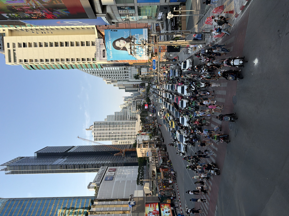 An elevated daytime photograph presents a bustling urban intersection in Bangkok, Thailand, specifically identified as the Asok intersection on Sukhumvit Road, with the Terminal 21 shopping mall prominently visible on the left. The foreground and midground are dominated by a wide, multi-lane street experiencing heavy traffic congestion, with vehicles, predominantly cars, taxis, and a significant number of motorcycles, appearing stationary or moving slowly. A large red and white striped zebra crossing spans the width of the visible road, densely populated by dozens of pedestrians navigating in various directions, some carrying bags or pushing small trolleys. Numerous motorcyclists, many wearing helmets and potentially working as delivery or ride-sharing drivers, are present on their bikes amidst the pedestrian traffic and in front of the car lanes.
The left side of the scene is anchored by modern high-rise buildings, including a tall glass skyscraper (G Tower) and the multi-story Terminal 21 shopping mall below it. The right side features additional tall buildings, some with distinctive architectural elements, adorned with large digital billboards; one notably displays a woman's face advertising "Bangkok Clinic," and another shows "Citi." The street extends into the background, revealing a dense urban canyon of further high-rise structures and a visible construction crane. The sky overhead is clear and blue, indicating fair weather. The overall scene depicts a vibrant commercial district characterized by intense vehicular and pedestrian activity.