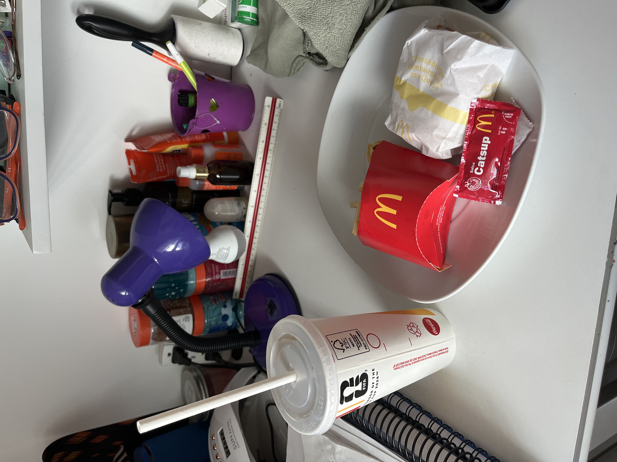 The image captures a cluttered white tabletop in what appears to be a personal space in San Bernardino Tlaxcalancingo, Mexico. The scene is dominated by a McDonald's fast-food meal. A white bowl contains a McDonald's cheeseburger, still in its wrapper labeled "Cheeseburger Hamburguesa con Queso," alongside a red carton of french fries. A red McDonald's ketchup packet, marked "Catsup M," "Cont. Net 8g," and "CORTE AQUI," is also inside the bowl.
Next to the meal, a white Coca-Cola cup with a plastic lid and straw is visible. The cup features prominent branding for the "FIFA WORLD CUP 2026™" and includes text such as "Hecho en Mexico Printed in Mexico" and "Coca-Cola es una marca registrada de The Coca-Cola Company," along with "FSC MIXTO FSC C178893."
The rest of the table is occupied by various personal items, suggesting a desk or nightstand setup. These include multiple bottles of toiletries (shampoos, lotions, sprays), a purple flexible desk lamp, a lint roller with a black handle, and a purple container holding pens or brushes. Several pairs of eyeglasses are visible on the far left. A ruler with red and white markings lies among the items, and a green towel or cloth is partially seen. Towards the bottom, a portion of a spiral-bound notebook and a small white electronic device displaying "SUN" and "2006" are present. The image portrays a casual, everyday setting, possibly after or during a quick meal.