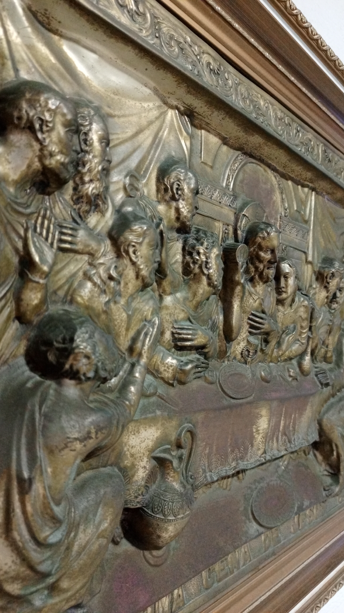 The image shows a bronze-colored relief sculpture depicting the Last Supper. Figures, presumably Jesus and the apostles, are seated at a table with plates and a pitcher. The sculpture is highly detailed, with intricate folds in the clothing and expressive faces. The background is textured and suggests a draped fabric. The scene is framed by an ornate border. The location in the city of Los Robles, Venezuela, is not discernible from the visual information.