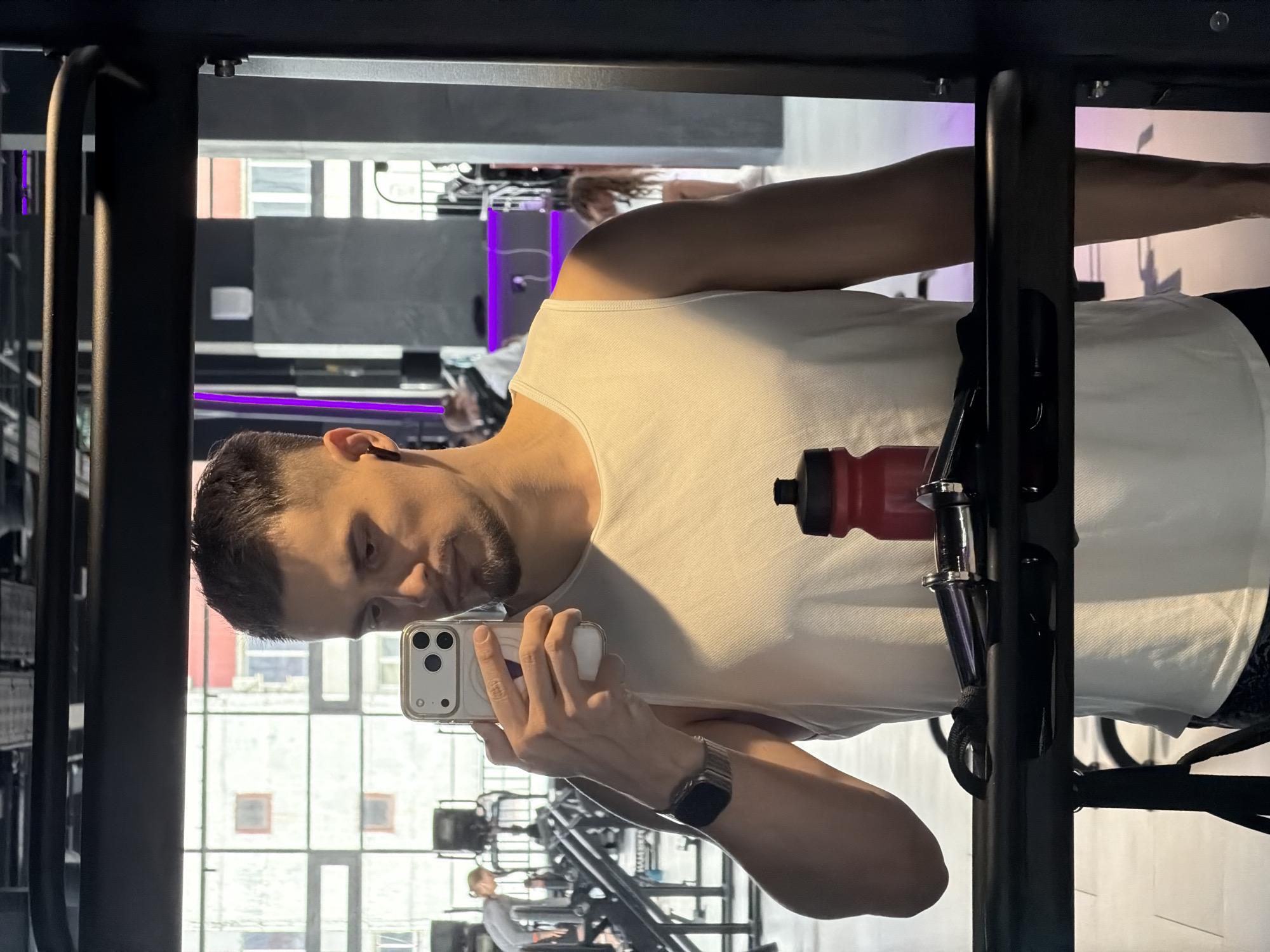 The image shows a man taking a selfie in a gym, possibly in Krasnoyarsk, Russia, as suggested by the architectural style of the building visible through the windows. He is wearing a white tank top and a watch, holding a smartphone in his left hand to capture the image. A red water bottle is visible, attached to a weightlifting machine. Gym equipment and other individuals exercising are seen in the background. The overall environment suggests a well-lit, active fitness facility.