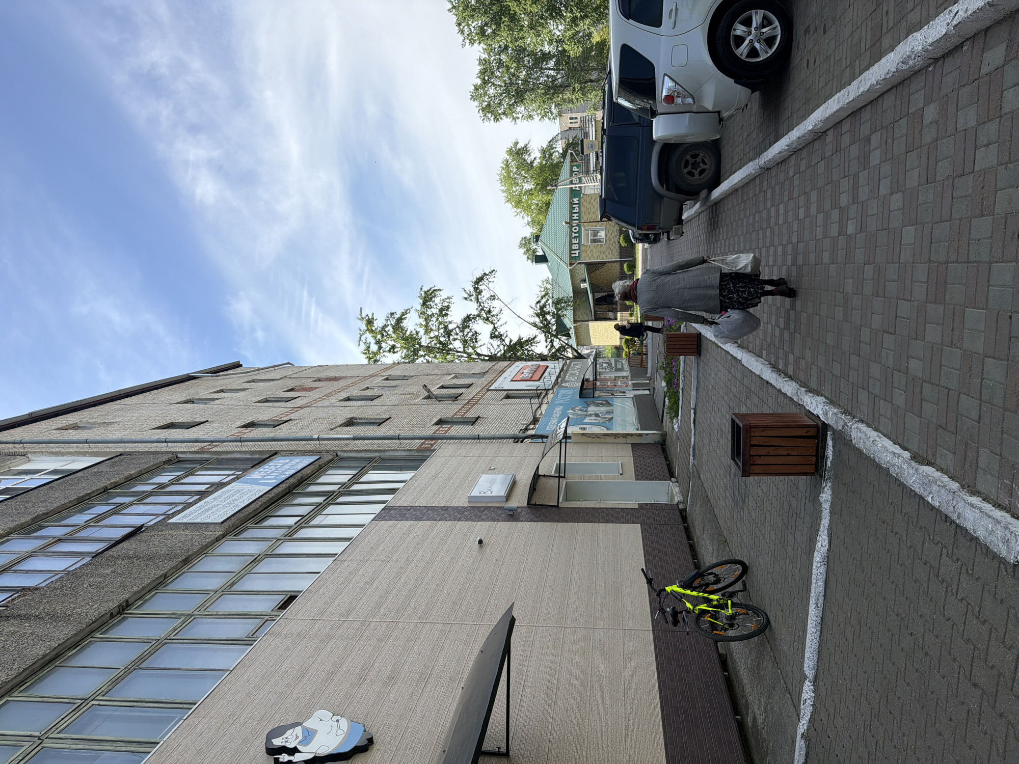 The image depicts an outdoor scene in what appears to be a city in Russia, likely Birobidzhan. A multi-story building with rows of windows dominates the left side of the frame. A sidewalk runs alongside the building, paved with dark bricks and bordered by a white line. A yellow bicycle is leaning against the building. An elderly woman walks along the sidewalk, carrying bags. Several parked cars are visible on the right side of the frame. Further down the street, there is a small building with a green roof and signage written in Russian. The sky is blue with scattered clouds.