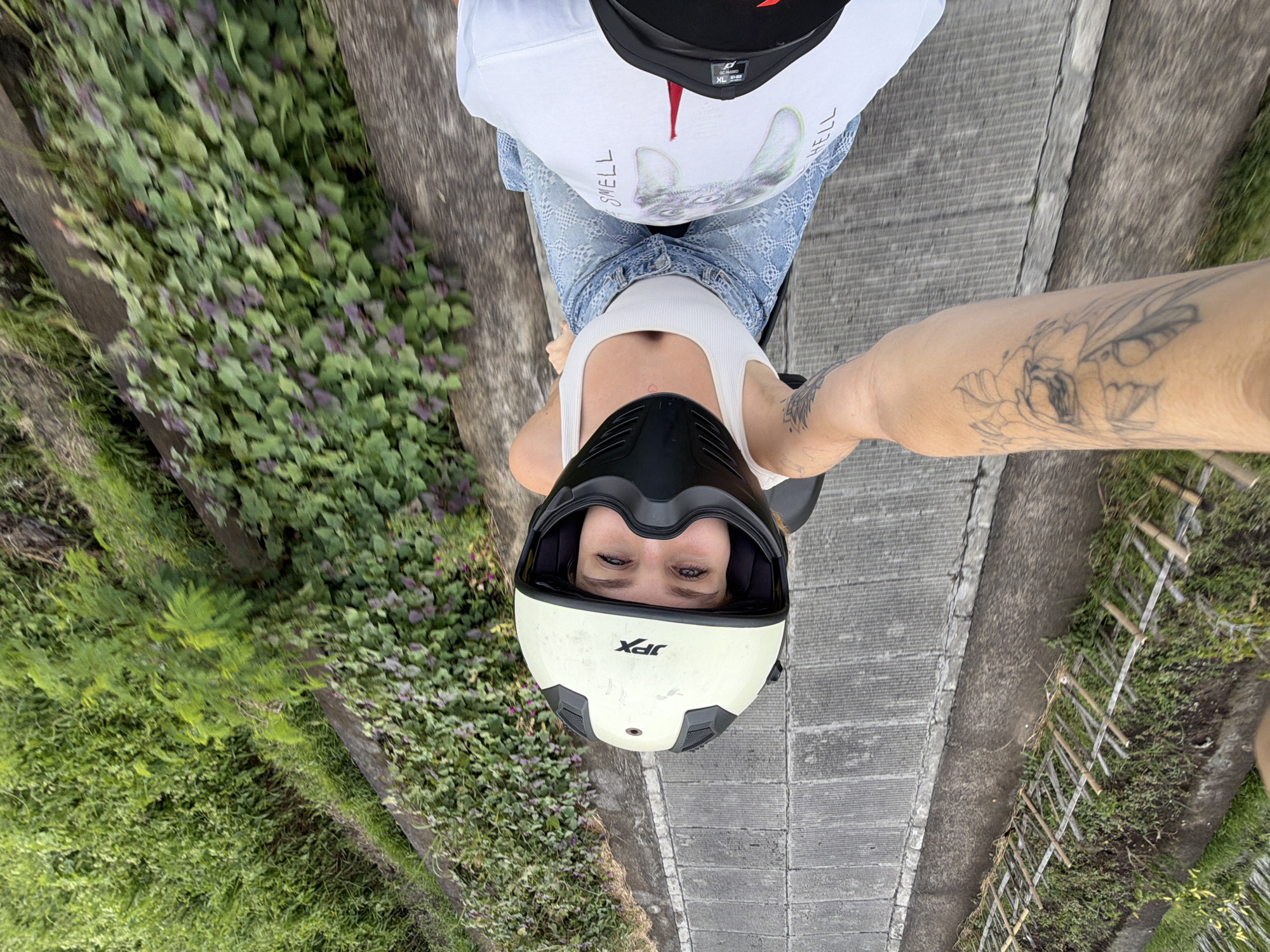 In North Kuta, Indonesia, a low-angle selfie captures two individuals seated on a scooter, moving along a paved path bordered by lush green vegetation. The person in the foreground, wearing a white tank top and distressed denim jeans, is wearing a cream-colored motorcycle helmet with black accents and a black face shield. Their gaze is directed towards the camera, with visible makeup on their eyes. Their arm, adorned with a tattoo of a flower, is extended, holding the camera. Behind them, another individual is visible wearing a white t-shirt with a graphic of a cartoonish dog and text that reads "SMELL HELL." The surrounding environment features concrete borders, a retaining wall, and dense foliage, including a mix of green leaves and some purple-hued plants.