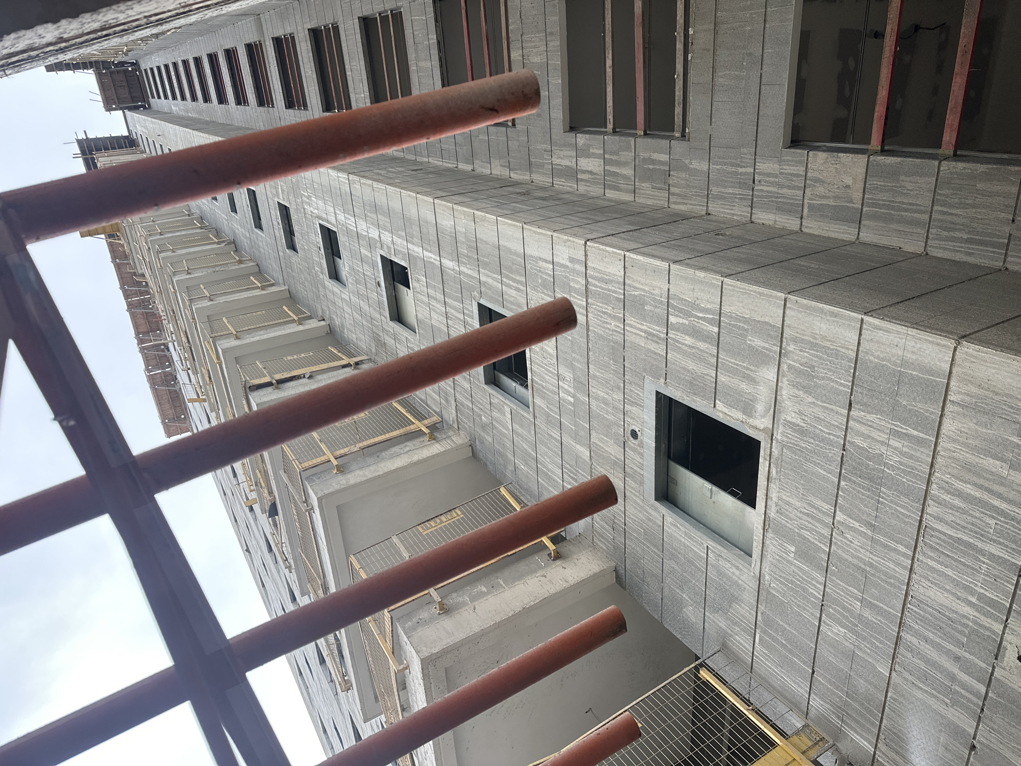 The image shows an exterior view of a tall, modern building under construction, viewed from below and looking upwards. The building's facade is made of large, grey stone tiles, with visible vertical streaks of water or dirt running down them, indicating recent weather or construction activity. Several rectangular windows or openings are visible, some of which are dark, suggesting they are either unfinished or have interior coverings. In some openings, red construction materials are visible.
The foreground is partially obscured by a diagonal scaffolding structure made of orange, cylindrical poles and horizontal metal bars. This scaffolding appears to be part of the construction process, possibly supporting temporary platforms or safety netting. The sky above is overcast.
The architectural style is contemporary, characterized by clean lines and geometric forms, typical of modern urban development. The building's height suggests it is likely a residential or commercial tower.
Given the style of construction and the general appearance, this image could be from a developing area in any modern city. Without specific visual cues unique to Haifa, it is not possible to definitively place this image in Haifa, Israel. However, such construction projects are common in rapidly developing urban centers.