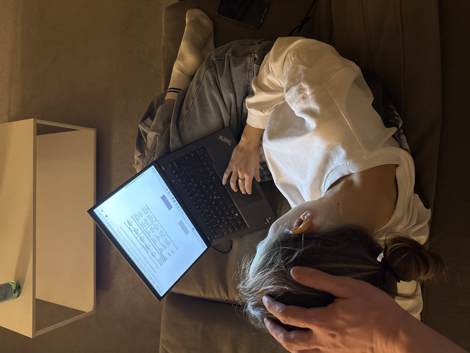 The image depicts an indoor, domestic scene. A person is positioned on a dark brown sofa, reclining with their head in the foreground. They wear a white long-sleeved shirt and light blue jeans. Their light brown hair is secured in a bun, and their right ear is visible. A black Lenovo ThinkPad laptop is resting on their lap, its screen illuminated and displaying text and tabular data, consistent with an online form or assessment. The person's left hand, adorned with a ring on the fourth finger, is positioned over the laptop's keyboard/trackpad area. A white sock covers their right foot, which appears to be encased in a cast or bandage. To the right of their leg, a black smartphone is partially visible on the sofa cushion. A second individual's left hand is visible, gently resting on the head of the person using the laptop, also wearing a ring on the fourth finger. In the background, a white, minimalist rectangular coffee table holds a green bottle. The wall behind the table is a plain, light brown or beige color. The specific location in the city of Venice, Italy, cannot be determined from the visual evidence present in the image.