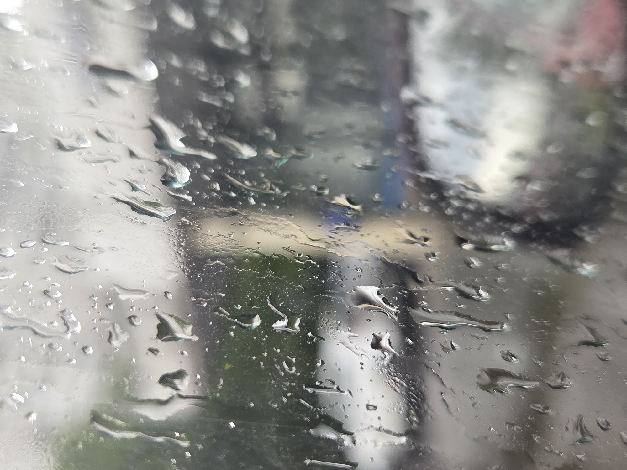 The image is a close-up, abstract shot of water droplets on a glass surface, likely a window or windshield. The droplets vary in size and shape, some appearing as small beads and others as larger, elongated streaks. They distort the view of the background, which is blurred and appears to be an outdoor scene. Hints of green suggest foliage, and muted grays and whites could indicate buildings or structures. The overall impression is that of a rainy or wet environment. The lighting suggests it is daytime, but the specific time is indeterminate. The location context of Moscow, Russia, provides a general setting, but no specific landmarks are visible. There is no discernible activity or event taking place; the focus is on the visual texture and abstract quality of the water on the glass. No text is visible in the image.