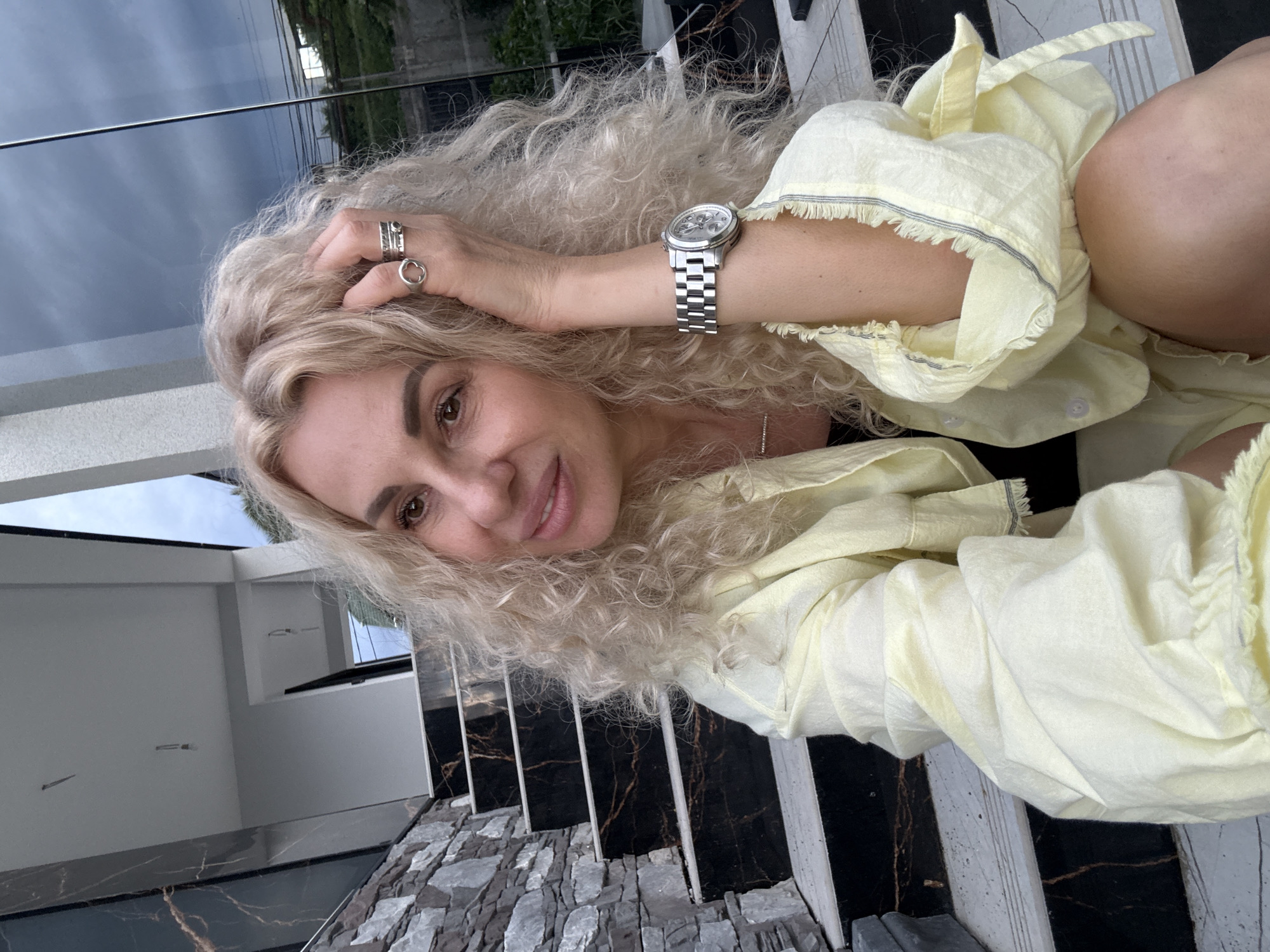 A blonde woman with curly hair and a yellow shirt sits on a staircase outside a modern building. The staircase has black marble steps with gold veins, and the wall to her left is made of gray stone. She is wearing rings and a watch, and her hand is resting on her head. The background shows a glass facade of the building, reflecting the sky and some greenery. The location is identified as Baranovka, Russia.