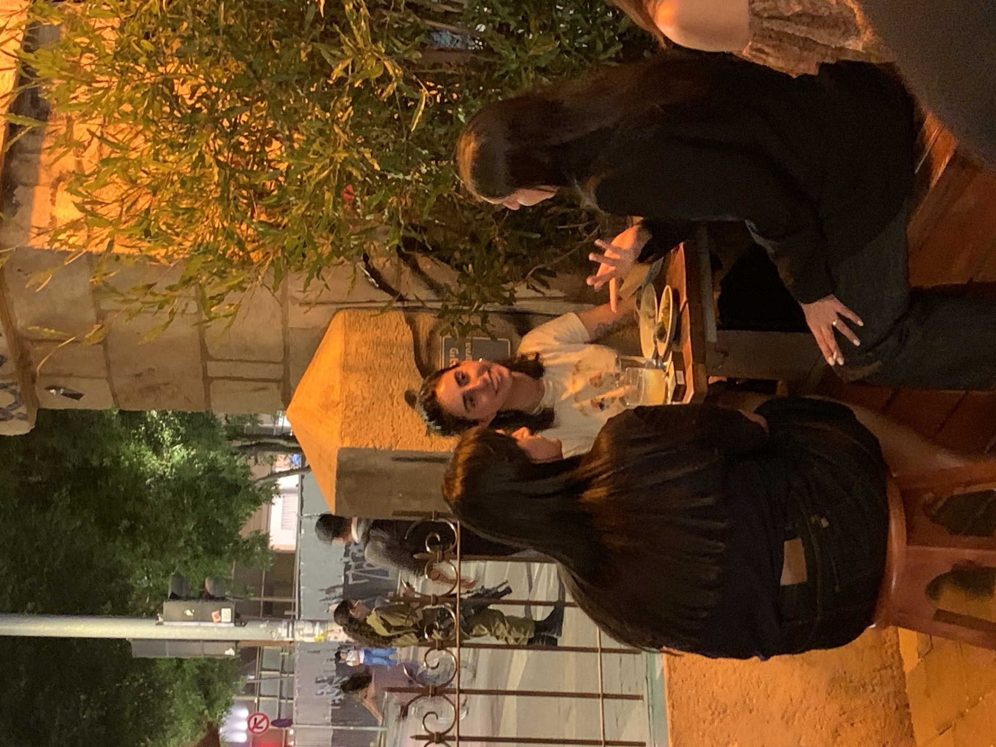 The image shows a group of people sitting at an outdoor cafe table in Tel Aviv, Israel. Two women are facing each other across the table, engaged in conversation. A third woman sits to the side, facing away from the camera. In the background, through a metal fence, two individuals in what appear to be military uniforms and carrying weapons are standing. Other pedestrians are visible behind the fence. The setting appears to be a restaurant patio with tables and greenery.