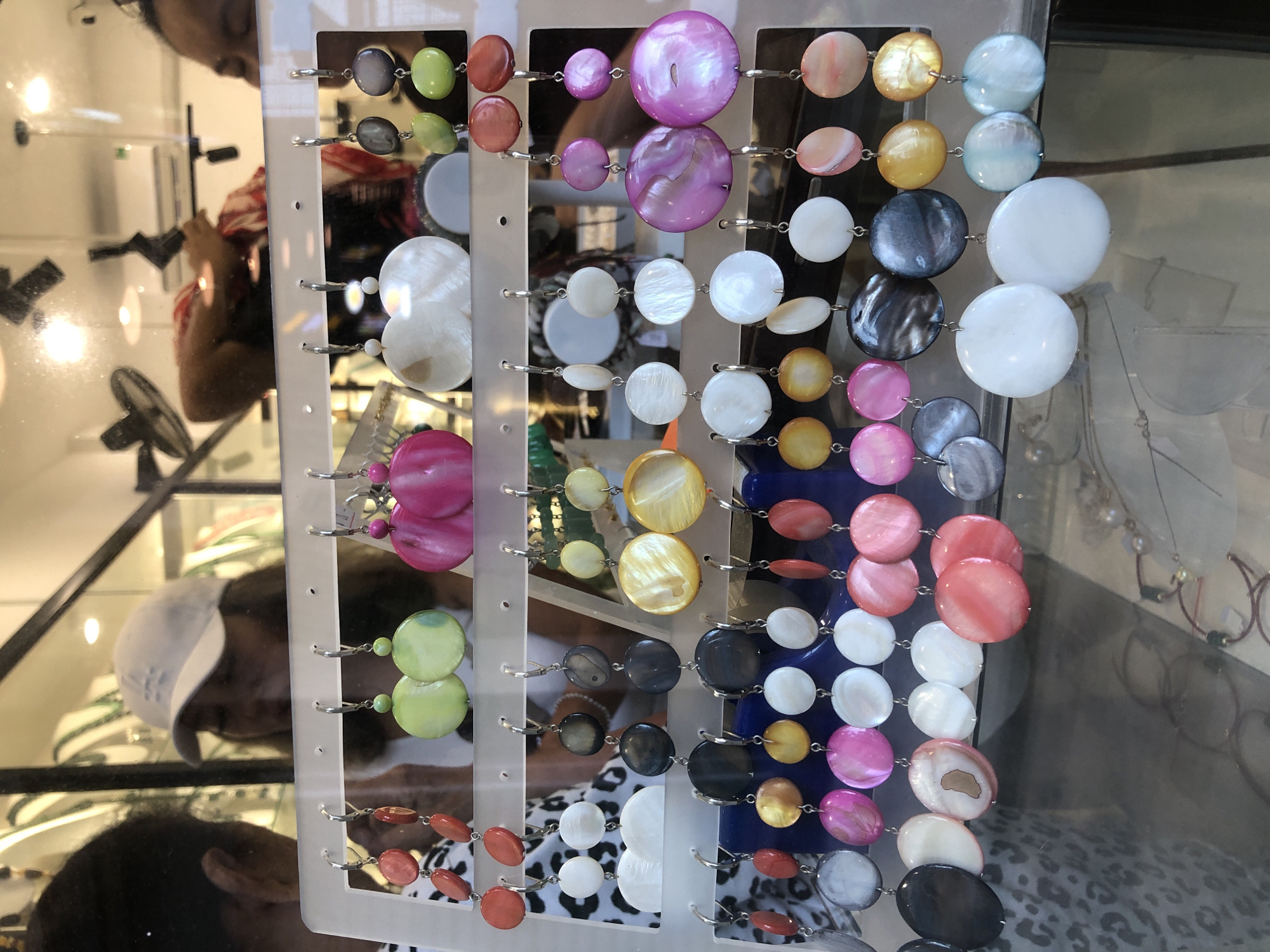 The image shows a display of colorful shell earrings at a market stall in Da Nang, Vietnam. The earrings are arranged in rows on a white stand, with various shapes and sizes, including round, oval, and fan-shaped designs. The colors range from light green and yellow to pink, purple, and white. In the background, reflections show people browsing and looking at the merchandise, suggesting a bustling commercial environment. The lighting appears to be artificial, possibly from indoor lighting or streetlights, casting a warm glow on the earrings.