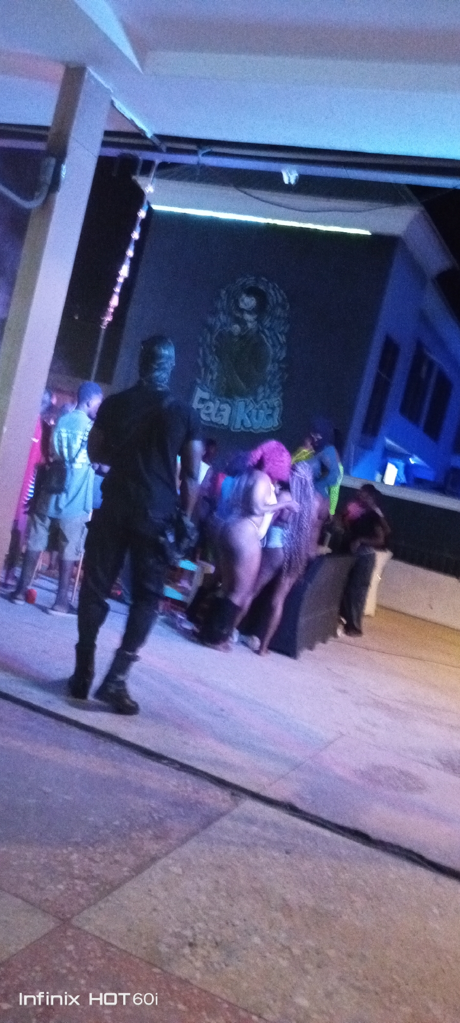 The scene is an outdoor gathering in Adankolo, Nigeria, based on the "Fela Kuti" sign, possibly at a bar or club. People are interacting, including individuals dancing or mingling. One figure in dark clothing faces away from the camera and toward the crowd. The environment includes stage lighting, a structure with a pole, and possibly a building in the background. The picture was taken with an Infinix HOT 60i phone.