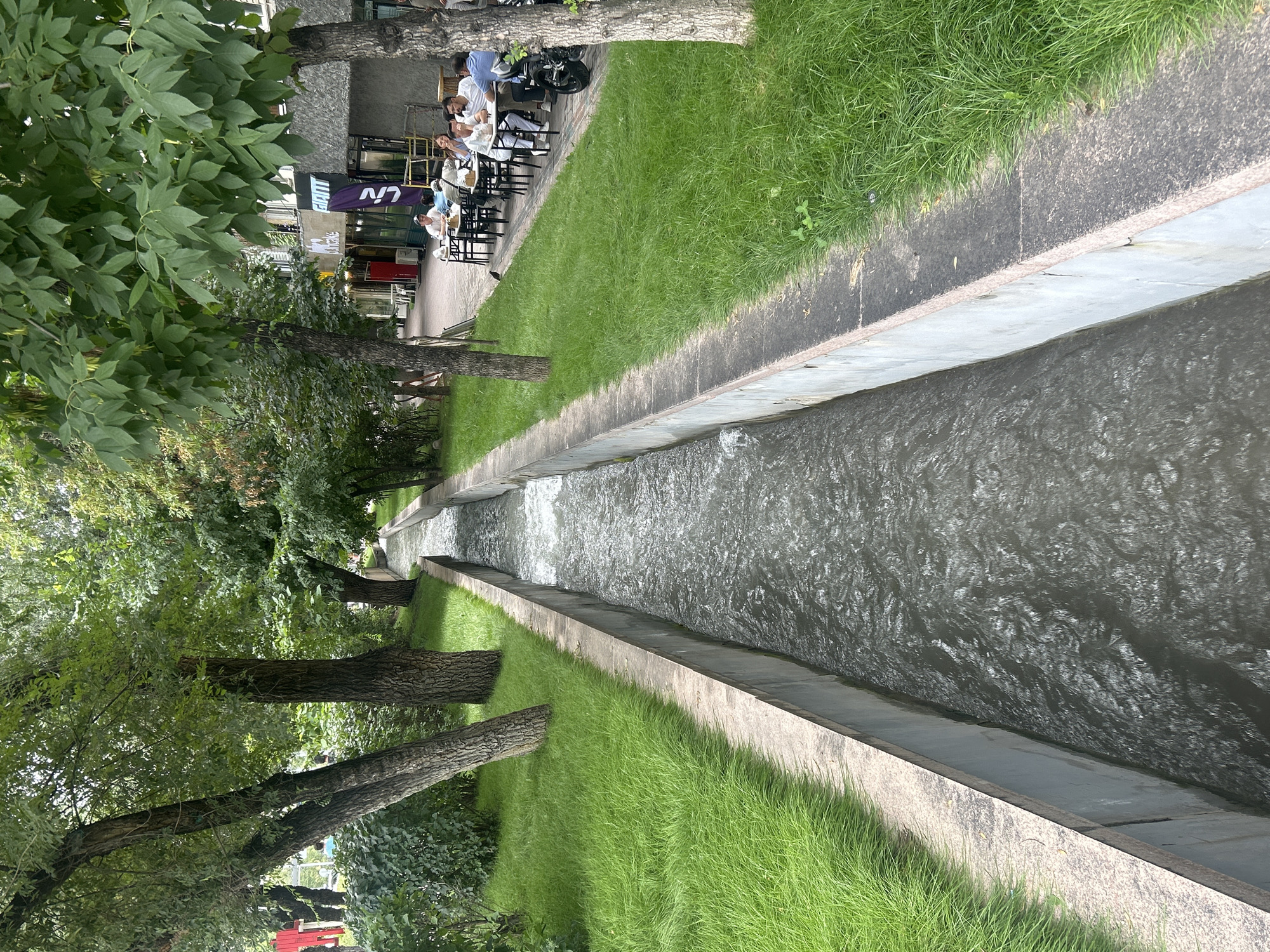 The image shows a tree-lined street in Almaty, Kazakhstan, with a concrete channel carrying flowing water down the center. Lush green grass flanks the channel on both sides. On the right side of the channel, a sidewalk runs alongside several shops and cafes. People are seated at tables outside one of the cafes, some eating and others conversing. A motorcycle is parked near the sidewalk. The buildings on the right appear to be commercial establishments with signage. The trees are mature with dense foliage, providing shade. The overall scene depicts a pleasant urban environment with a focus on greenery and outdoor amenities.