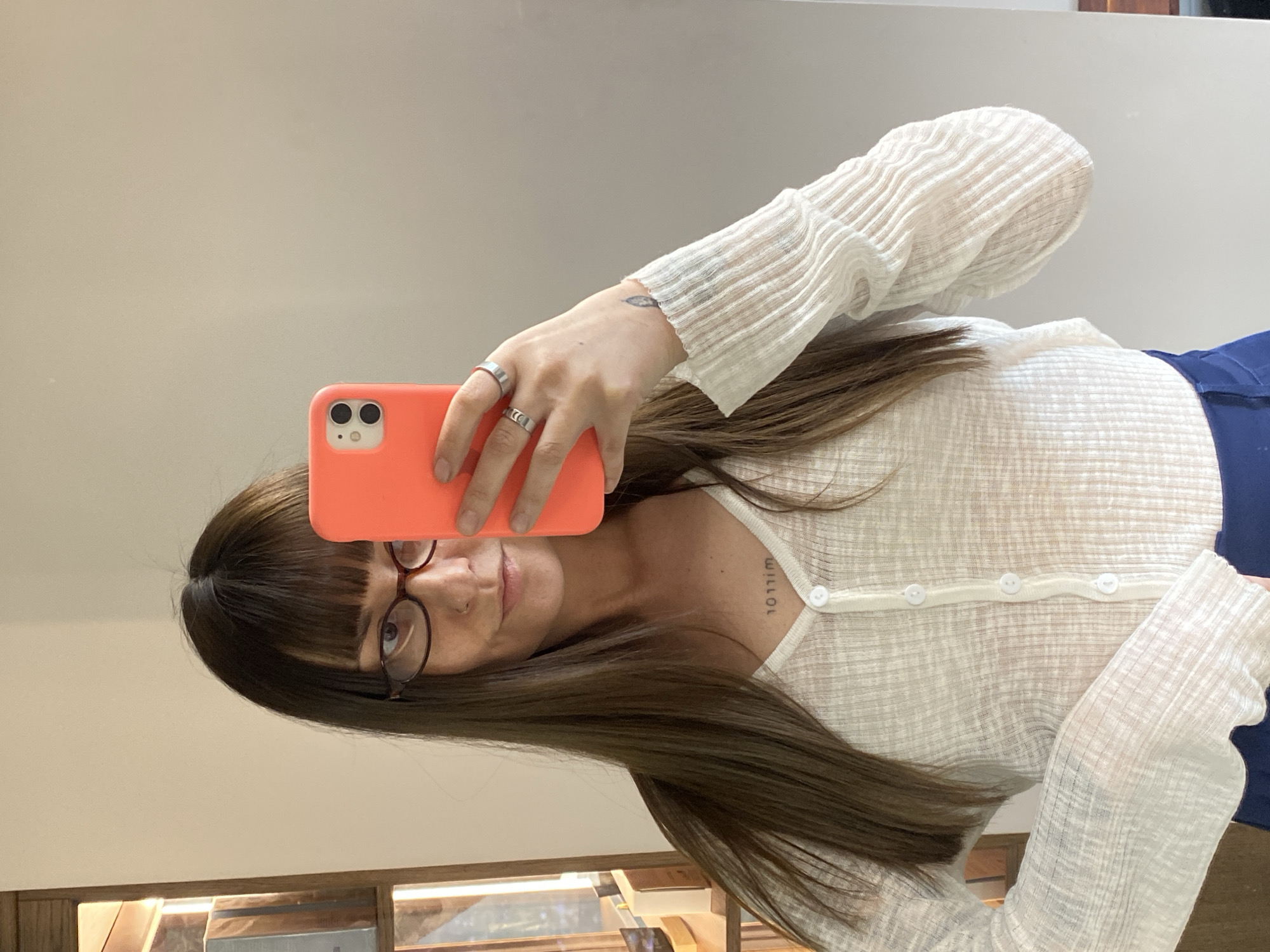 A woman with long, straight brown hair and bangs takes a mirror selfie, holding a smartphone with a bright orange case that partially covers her face. She wears round-framed glasses and looks towards the camera with a neutral expression.
She is dressed in a sheer, light-colored, long-sleeved top or cardigan featuring a textured or ribbed fabric and a row of white buttons down the front. A dark blue garment, possibly pants or a skirt, is visible on her right side. On her left hand, she wears two silver rings, and a small, delicate tattoo, possibly a feather or leaf design, is visible on the back of her hand. Another tattoo appears on her collarbone, depicting text that reads "10111m" or "willol" (inverted from the viewer's perspective).
The background is a plain, light-colored surface, suggesting an indoor setting with a mirror. At the bottom of the frame, there are faint reflections or details that might indicate wooden shelving or furniture.
