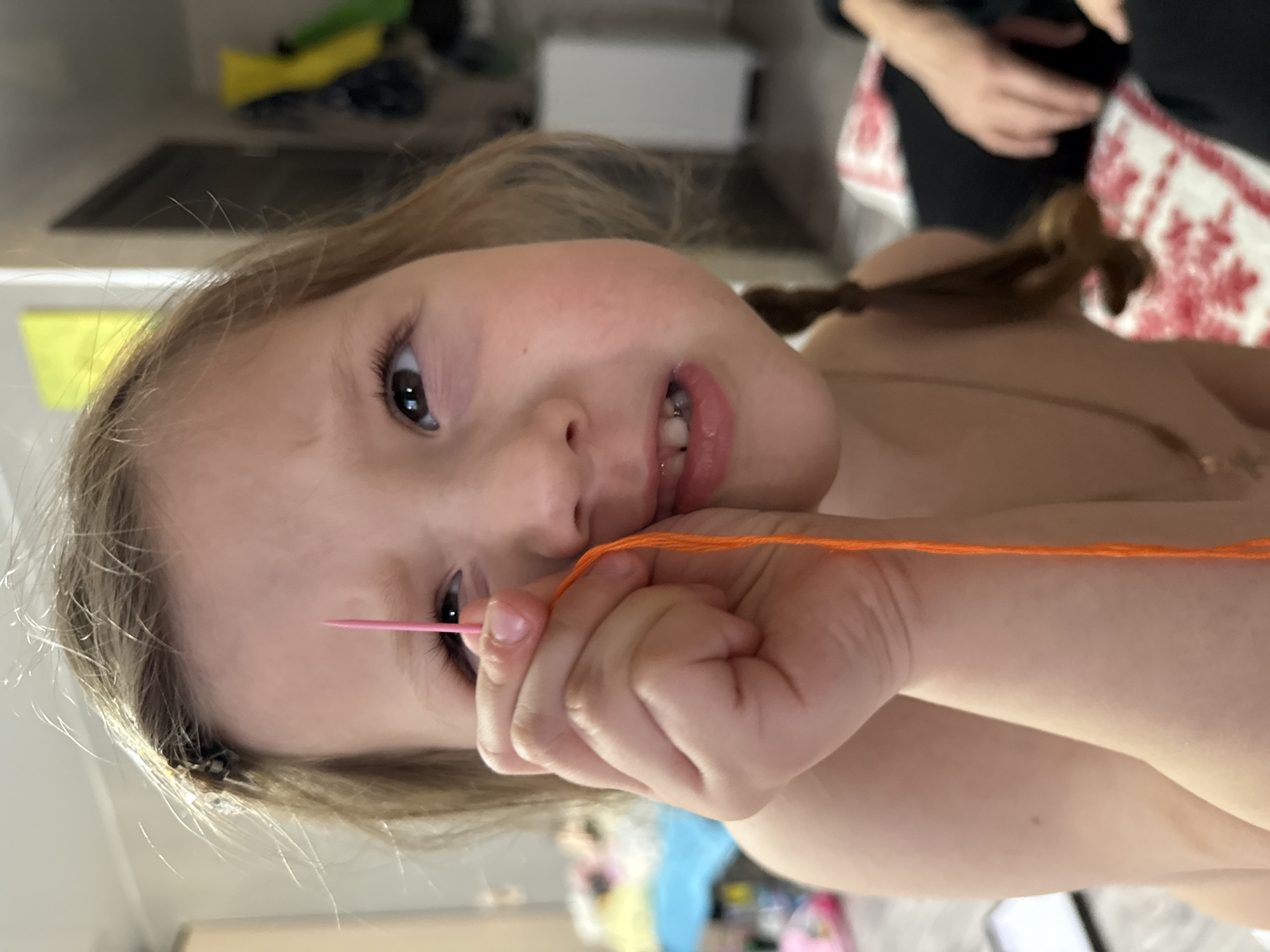 A young girl is depicted in a close-up, angled shot, holding a needle and orange thread. Her face is centered, with her eyes looking directly at the camera. Her blonde hair is partially pulled back in a braid. The background is blurred, showing a glimpse of a room with various objects and a person's arm in the upper right corner. The location is not evident from the image and therefore the location in Кипчак, Russia is not confirmed.