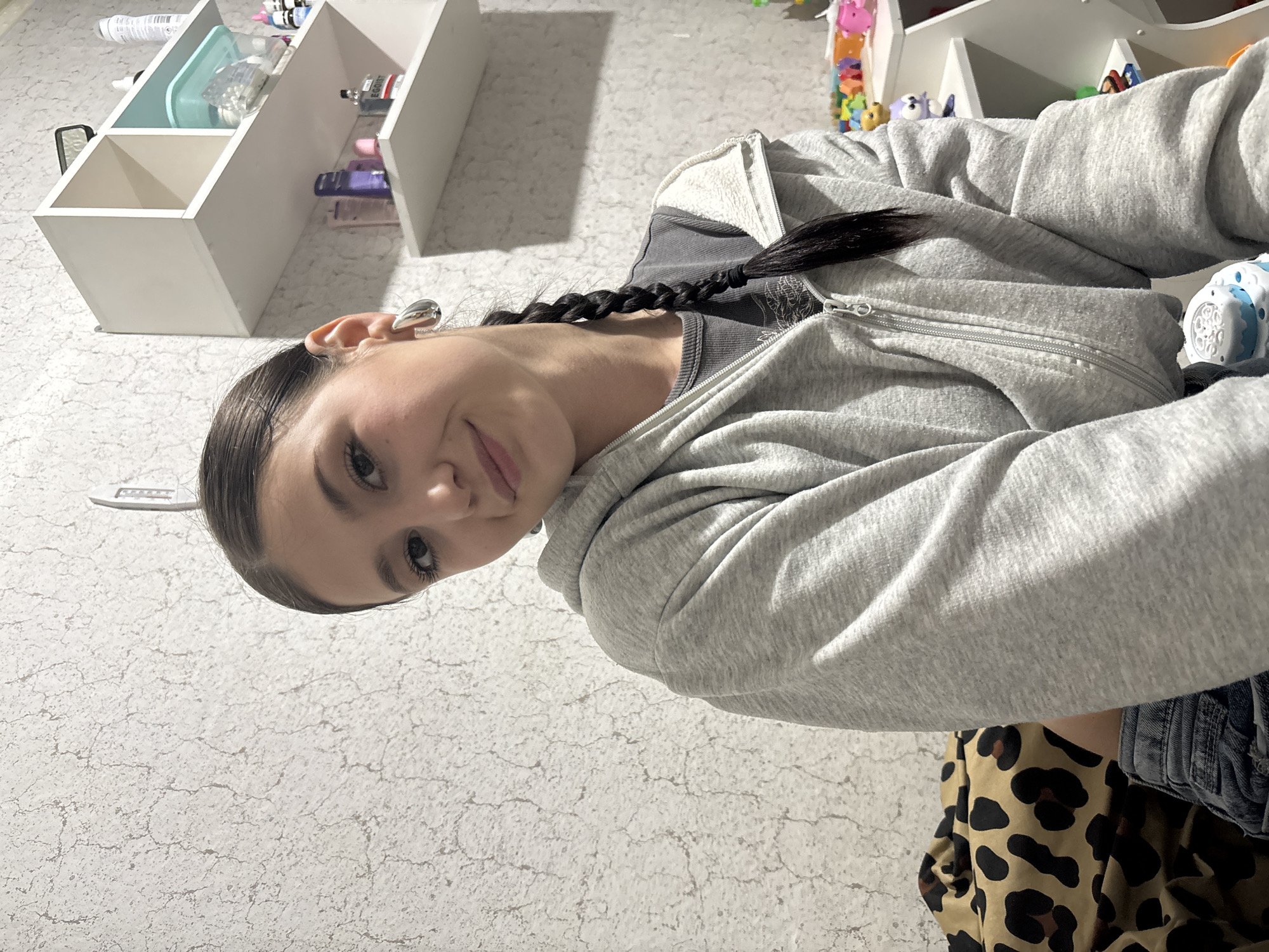 A young woman with dark hair styled in a braid is facing the camera. She is wearing a gray zip-up hoodie over a dark gray shirt, and a leopard print top is visible. To the left of the woman is a white shelf with several compartments holding various items. The background is a mottled off-white color, likely a wall or floor. The location is not possible to determine from the given image.
