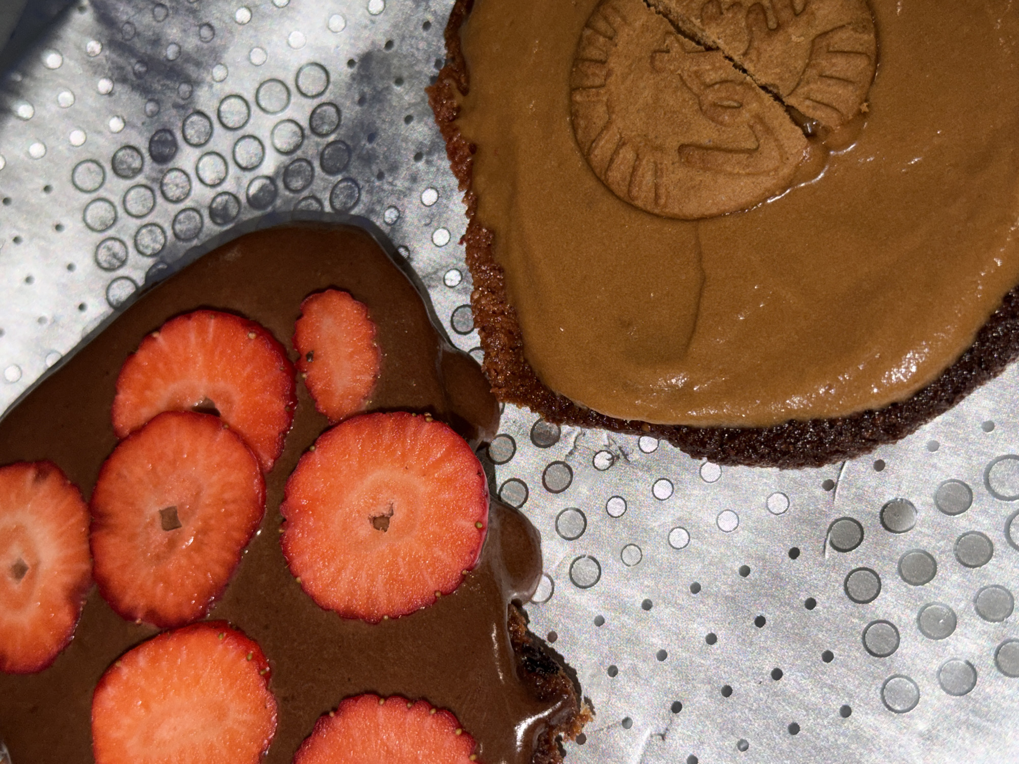 The image shows two desserts on a perforated metal surface. One dessert is covered in chocolate frosting and topped with sliced strawberries. The other dessert has a light brown frosting and a broken biscuit on top. The desserts are close together on the surface. The background is the perforated metal surface. The image does not contain information about the city of Varna, Bulgaria.
