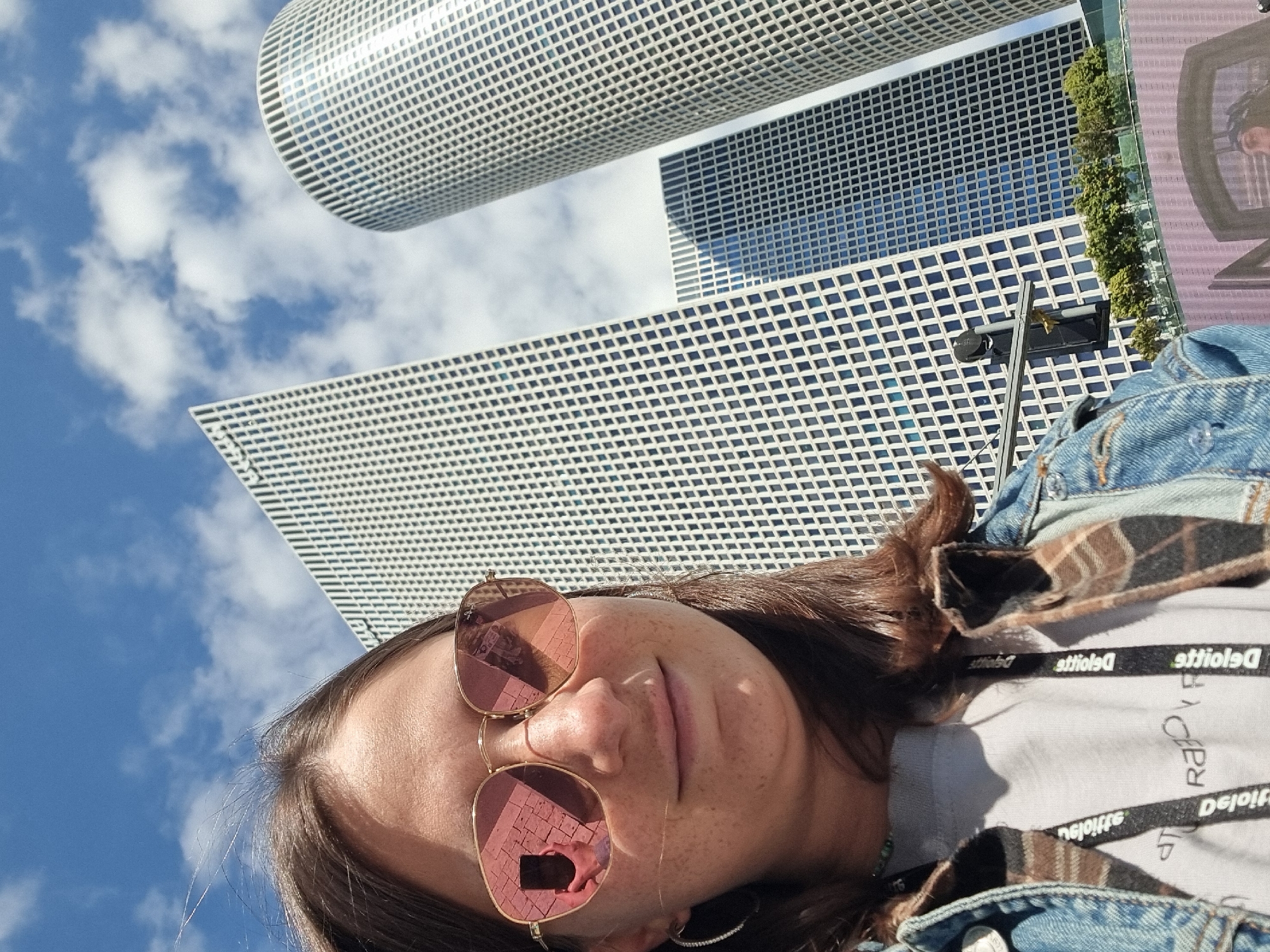 The image is a low-angle selfie taken in Tel Aviv, Israel, with the subject, a woman wearing rose-tinted sunglasses, filling the lower half of the frame. The reflection in her glasses reveals a partial view of the surrounding cityscape. Behind her, the modern architecture of Tel Aviv is prominently featured, including the Azrieli Center's circular tower and other high-rise buildings with grid-like facades. A blue sky with scattered clouds is visible. The woman is wearing a denim jacket over a light gray shirt, with a black lanyard bearing the text "Deloitte" around her neck.