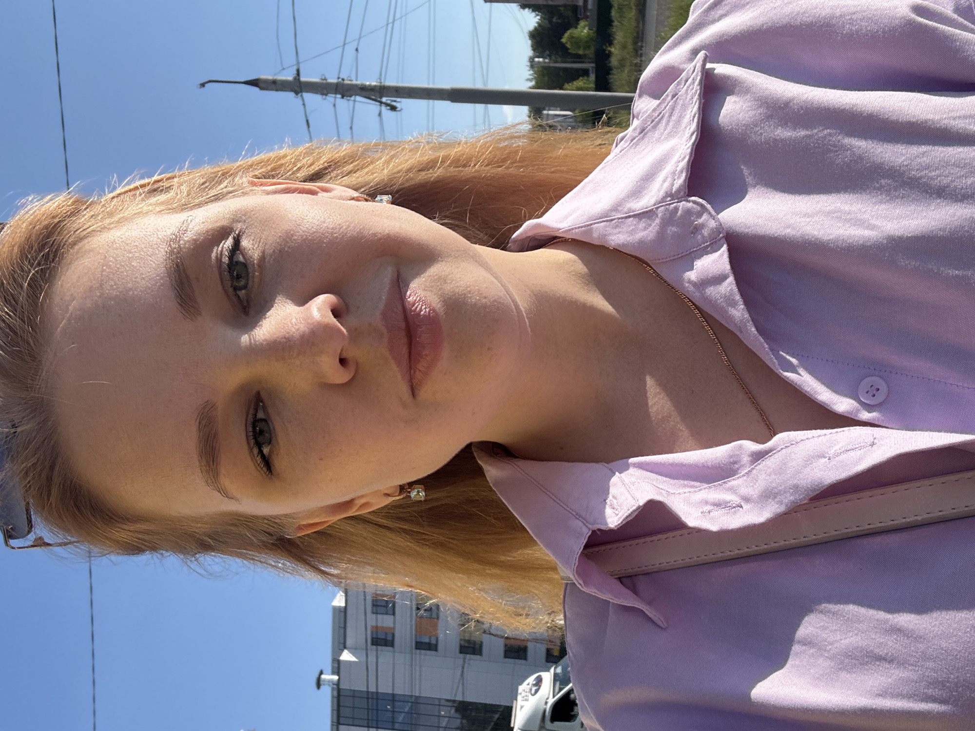 A close-up, selfie-style portrait of a woman with auburn hair is captured outdoors on a sunny day. She is wearing a light purple collared shirt and a thin gold necklace. A light-colored purse strap is visible across her shoulder. In the background, a modern apartment building and a utility pole with wires can be seen. The scene appears to be located in an urban or suburban area, possibly on a street or sidewalk, with a clear blue sky overhead. The specific location is not visually identifiable as Шебунино, Russia, from the image alone.