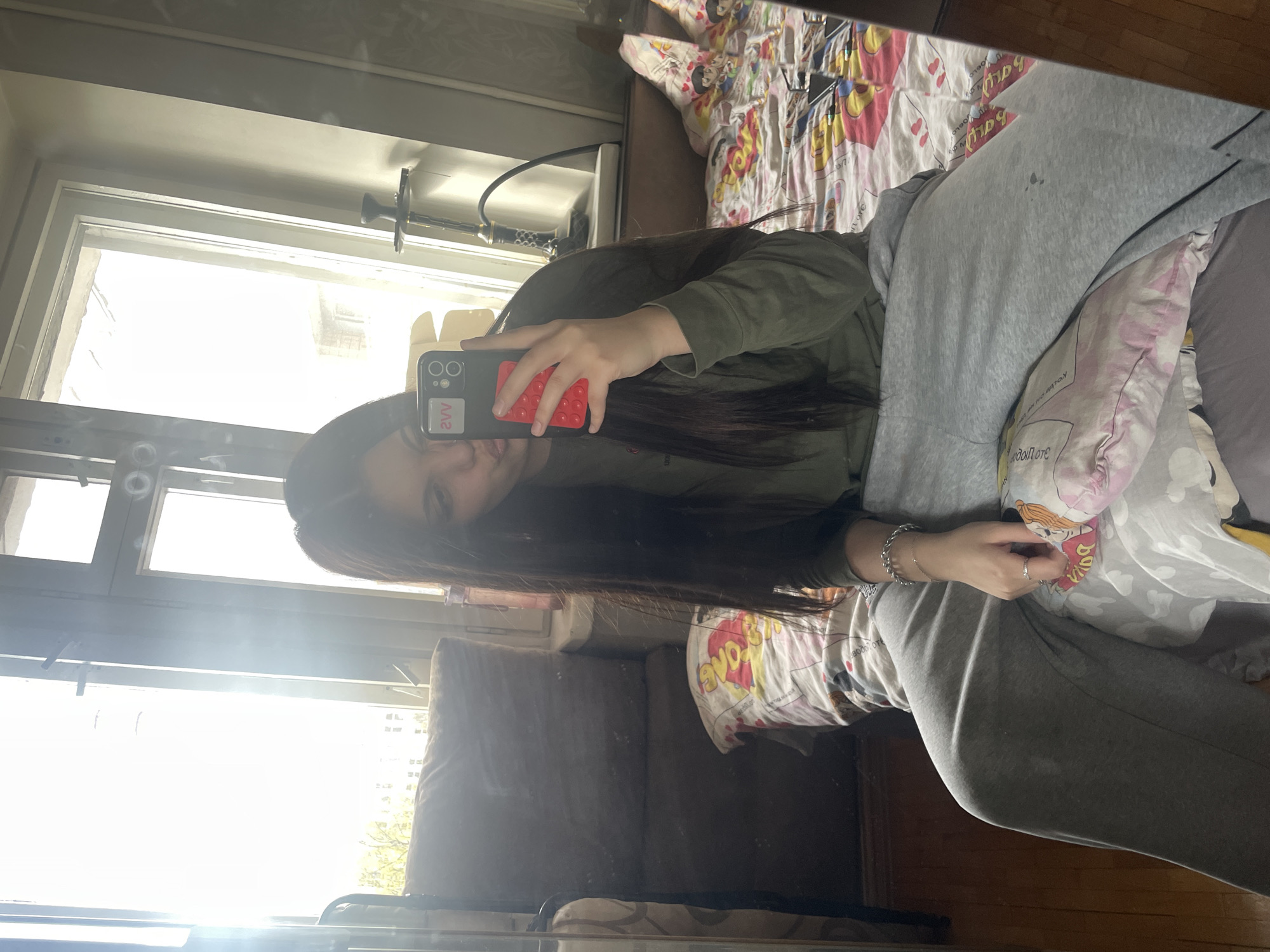 The image is a mirror selfie taken indoors, depicting a young person in a casual setting. The photograph is rotated 90 degrees clockwise from its intended upright orientation.
The main subject is a young individual with long, dark hair, wearing a dark green long-sleeved top and grey sweatpants. Their face is partially obscured by the smartphone they are holding up to take the picture. A silver chain bracelet adorns their left wrist, and a ring is visible on one finger. The phone is black with a distinctive red "pop-it" style case, and a white rectangular element on its back displays the mirrored text "2W" (which would be "W2" unmirrored).
The background reveals a room, likely a bedroom or living area. To the right of the person, a bed is made with colorful, patterned bedding featuring cartoon characters and various texts. Mirrored words visible on the bedding include "babg", "yovo", and "LOVE" next to a heart symbol on a white sheet with yellow and red motifs. Another pink and white patterned sheet shows additional mirrored text such as "yovo", "OTOLPOJ", and "YI".
A large window dominates the left side of the reflection, letting in very bright daylight, suggesting the photograph was taken during daytime, possibly sunny or very bright overcast. Adjacent to the window, a hookah or shisha pipe with a black hose and metal top is partially visible on a surface. A dark brown sofa or armchair is also present in the room. The overall scene captures a candid, everyday moment in an indoor space, consistent with a residence in Saint Petersburg, Russia.