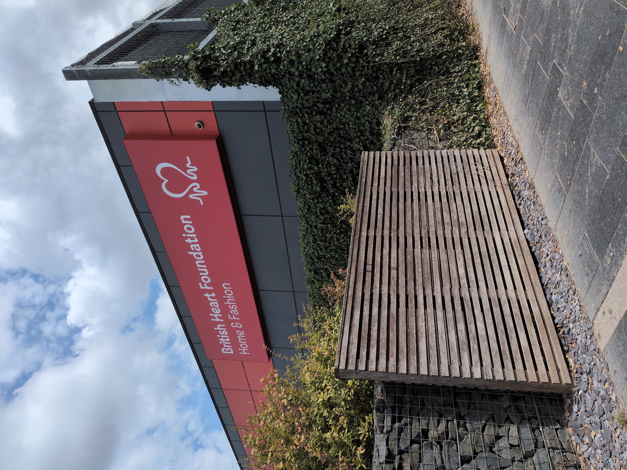 A British Heart Foundation Home & Fashion store sign is displayed on the side of a modern building with a grey facade and a red accent panel. The sign features white lettering and a white heart logo with an electrocardiogram line. To the right of the building, dense green ivy covers the wall, and a metal fence is visible above the ivy. In the foreground, a weathered wooden slatted screen is positioned in front of a gabion wall filled with stones. The ground consists of a paved walkway with rectangular grey tiles, bordered by small grey stones and fallen leaves. The sky above is partly cloudy with white cumulus clouds.