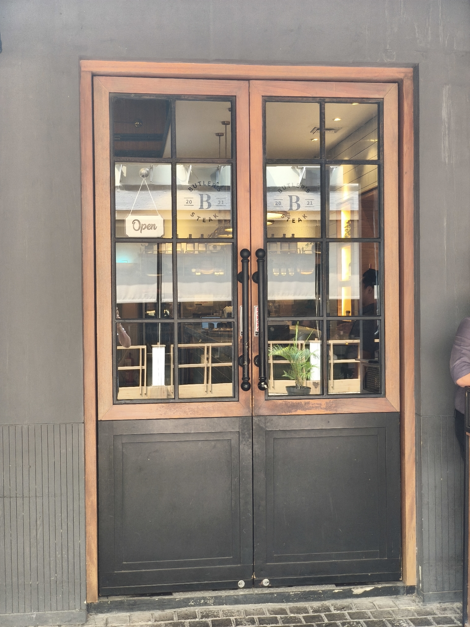 The image shows a storefront with double doors made of wood and black metal panels. The upper section of the doors consists of glass panes divided by a dark metal grid. A sign that says "Open" is hanging from one of the glass panes. Through the glass, a restaurant interior with tables and chairs is visible, along with some people. The exterior wall is dark gray and textured. The image is taken from a frontal perspective, at eye level. The location is the city of South Jakarta, Indonesia.