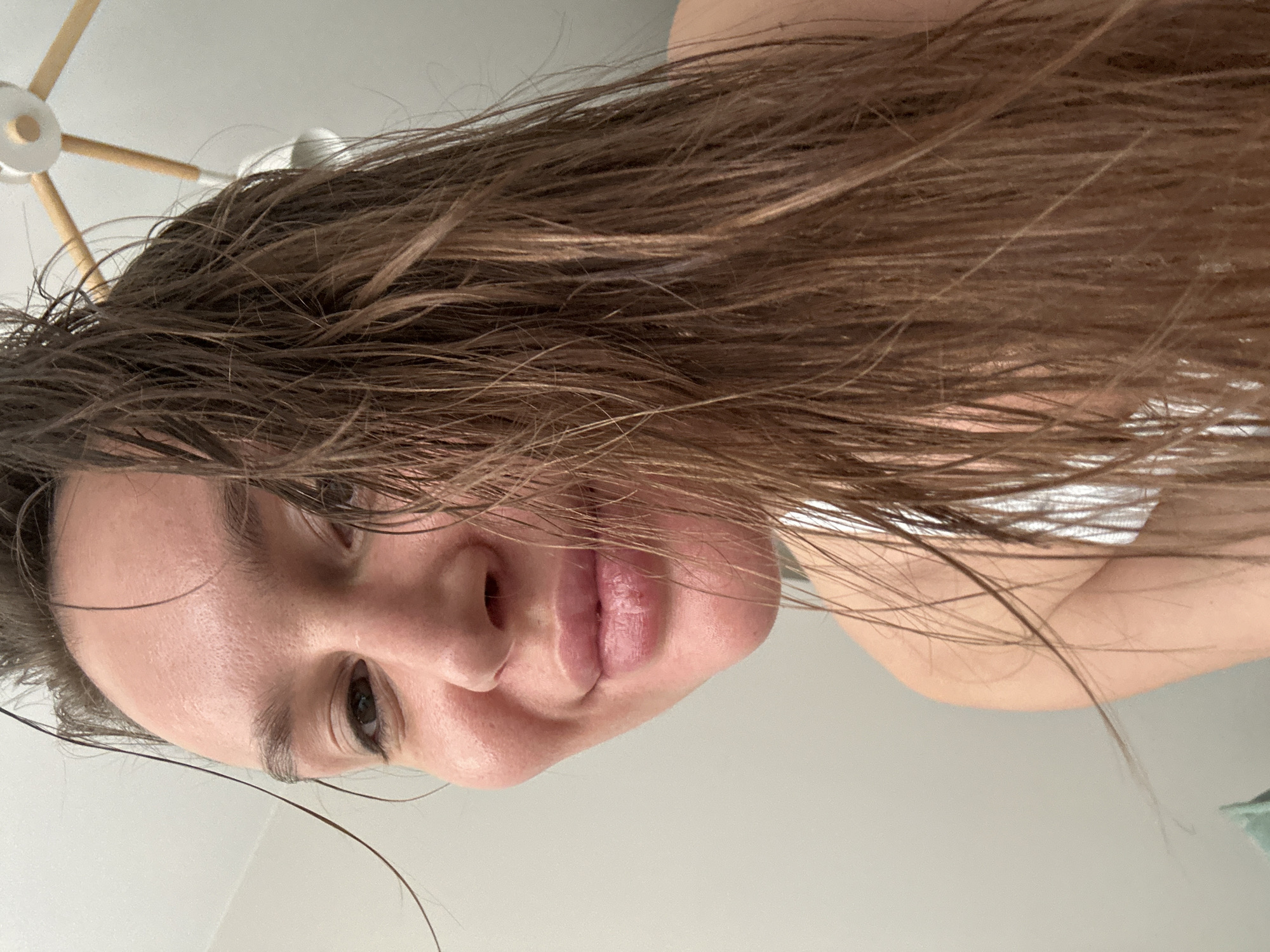 The image is a portrait-oriented close-up of a person, likely female, whose head is positioned on the left side of the frame, looking towards the camera. Their long, brown hair, appearing wet or damp, extends from their head upwards and across the majority of the top and right sections of the image, with individual strands visible.
The person's face is partially visible on the left. Their right eye, dark brown, is open and looks directly at the viewer, framed by a well-defined eyebrow. Below the eye, a portion of the nose is visible. The lips are closed, full, and a natural pink hue, appearing slightly glossy or damp. The skin on the visible part of the face seems smooth and has a subtle sheen, consistent with recent washing.
On the right side of the image, a small area of a light-colored shoulder or upper arm is visible, suggesting the person is wearing a light-colored garment, possibly white. A hint of a green item, likely clothing or fabric, is visible at the far right edge.
In the upper left corner, partially obscured by hair, is a modern-looking ceiling fixture with a white central component and several wooden spokes radiating outwards. The background throughout the rest of the image is a plain, light grey or white surface, likely a wall or ceiling.
The overall impression is an indoor setting, possibly a domestic environment like a bathroom or bedroom. The wet hair suggests the person might have recently showered or washed their hair. Their expression is neutral and direct. No text is visible in the image. The location context provided indicates this scene is in Moscow, Russia.