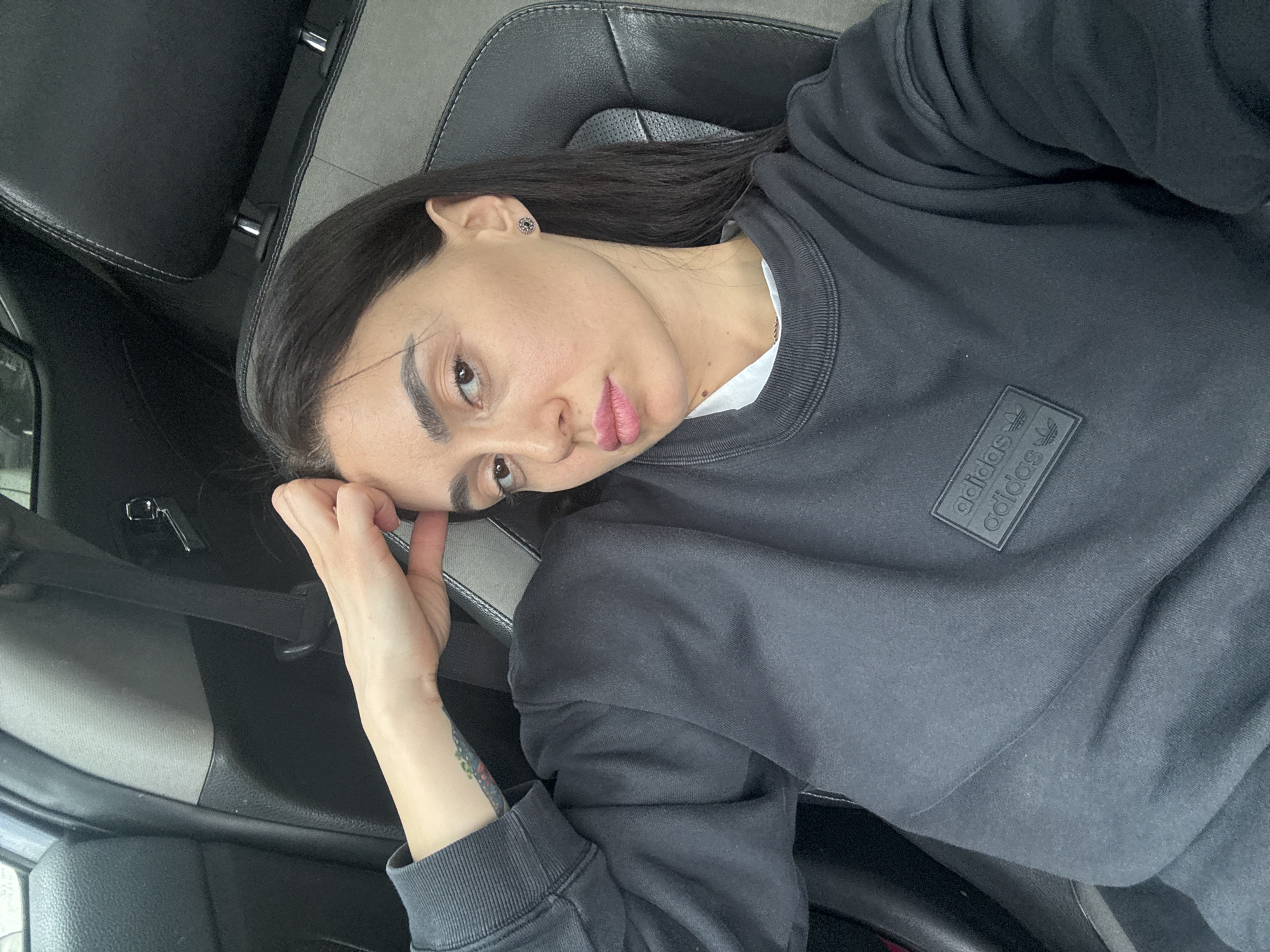 A single adult female is depicted in a medium close-up, seated inside a vehicle. She has dark hair, fair skin, and brown eyes, looking directly at the camera. Her lips are slightly parted, exhibiting pink lipstick. A small, dark, circular earring is present in her right earlobe. She wears a black long-sleeved crewneck sweatshirt with two rectangular black patches featuring the "adidas" text and trefoil logo on the left chest area. A white collar or undershirt is visible at her neck. Her left arm is raised, with the hand resting against her left temple/forehead. A multicolored tattoo is partially visible on the exterior of her left forearm, near the wrist.
The immediate environment is the interior of a car, characterized by black and grey upholstery on the seats and headrests. A black seatbelt is visible diagonally across her right shoulder. The vehicle's interior components, including door paneling and headrests, are dark grey and black. There are no discernible external visual cues in the image to determine a specific location within the city of Tbilisi, Georgia.