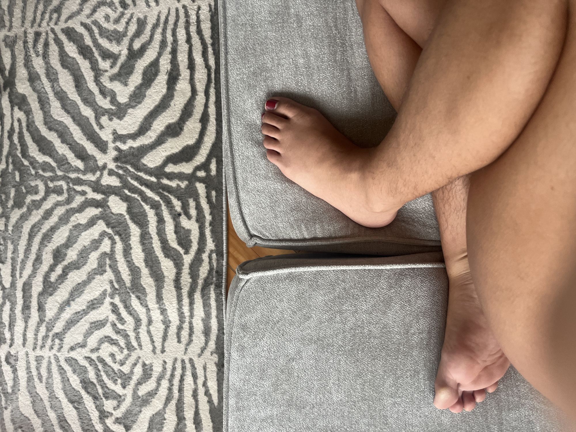 The image shows a partial view of a person lying on a gray upholstered surface, possibly a sofa or sectional, next to a gray and white zebra-patterned rug. The person's legs are crossed, with one foot resting on the cushion. The toenail of the big toe on that foot is painted red. The scene is indoors, with a glimpse of a wooden floor visible between the cushions. The location is not determinable from the image.