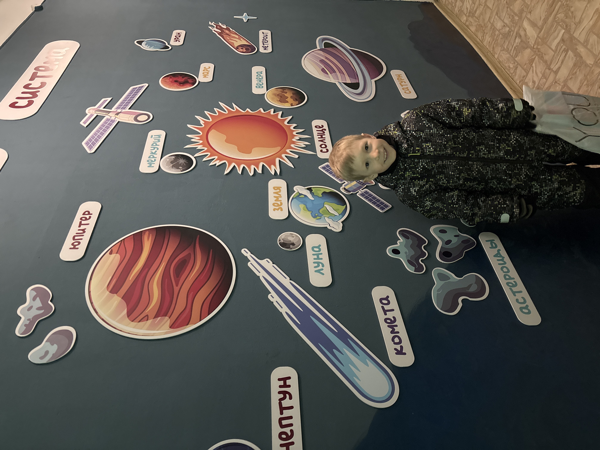 The image shows a young boy standing next to a wall decorated with a solar system theme. The planets and other celestial objects are depicted as cartoonish stickers. The boy is wearing a dark patterned winter jacket. The planets and objects visible include the Sun, Earth, Jupiter, Neptune, a comet, asteroids, the moon, and other planets. The text on the wall is in Russian. The location is Chaikovsky, Russia.