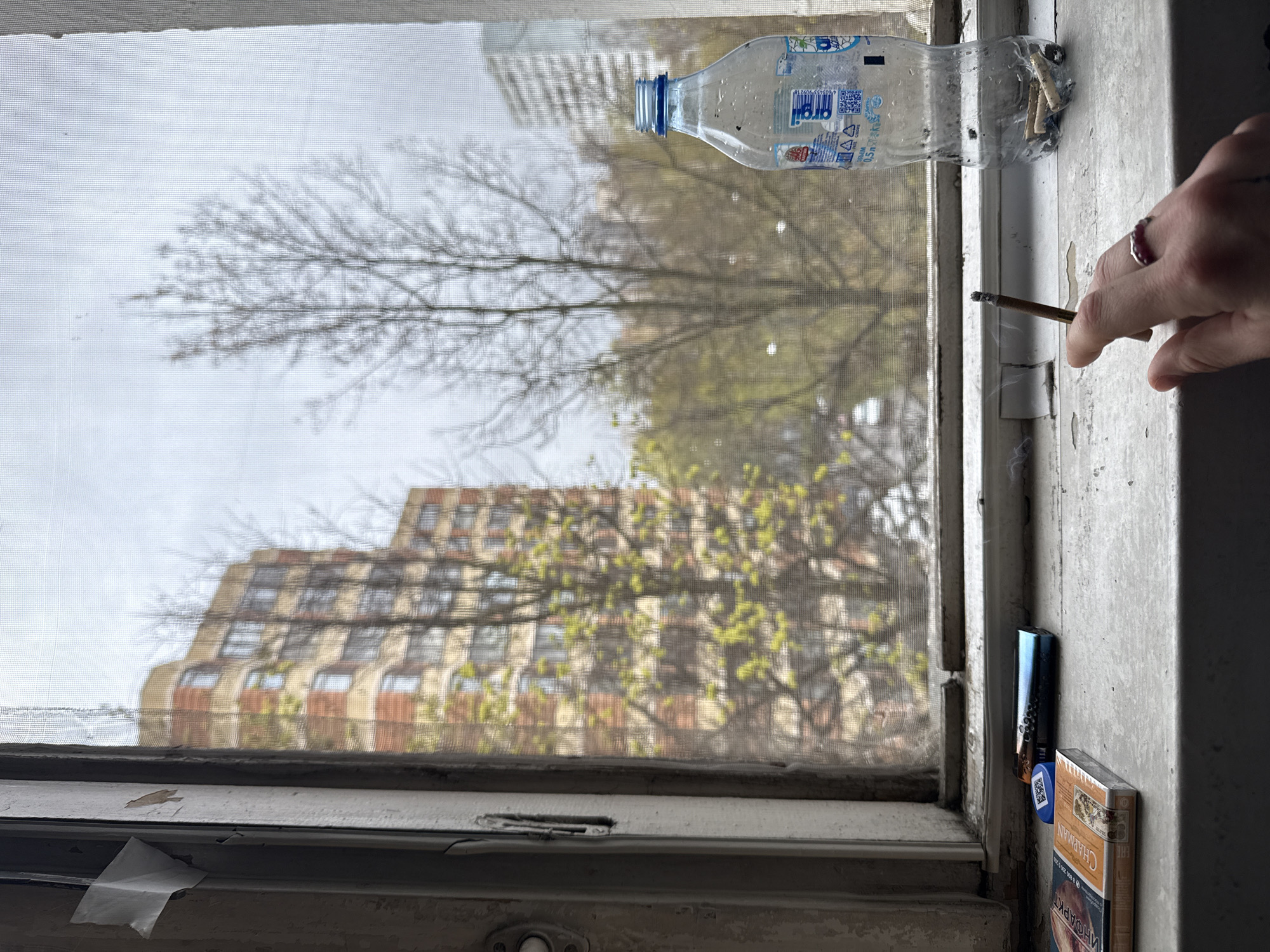 The image captures an indoor perspective, looking out through a mesh window screen at an urban scene, with a slight vertical rotation. In the immediate foreground on the right, a person's hand, adorned with a ring featuring a red or purple stone, holds a lit cigarette or joint, from which a thin wisp of smoke rises.
The windowsill, appearing somewhat worn with peeling paint, holds several objects. A clear plastic water bottle, cut horizontally and repurposed as an ashtray, contains multiple cigarette butts. The bottle's label displays Cyrillic text, including "0.5л" (0.5 liters) and what appears to be a brand name like "OROLA." Next to it, a "CHAPMAN" brand cigarette pack with Cyrillic text, including a health warning like "ИНОФАРКТ" (Infarct), rests beside a small, blue and black electronic device, possibly a vape or lighter. A piece of white tape is visible on the lower window frame.
Beyond the window screen, the outdoor scene is diffused but discernible. The sky is overcast or cloudy, indicating daytime. Trees, some still bare and others showing nascent green leaves, suggest early spring. A large, multi-story building with numerous windows and a distinctive architectural style stands prominently. Faintly in the distance, a city skyline is visible.
The activity depicted is a person smoking by a window, likely for ventilation, and using a makeshift ashtray. The visible Cyrillic text on the bottle and cigarette pack, particularly the "CHAPMAN" brand which is Russian, strongly supports the given context of Moscow, Russia. The overall atmosphere conveys a lived-in, utilitarian environment.
