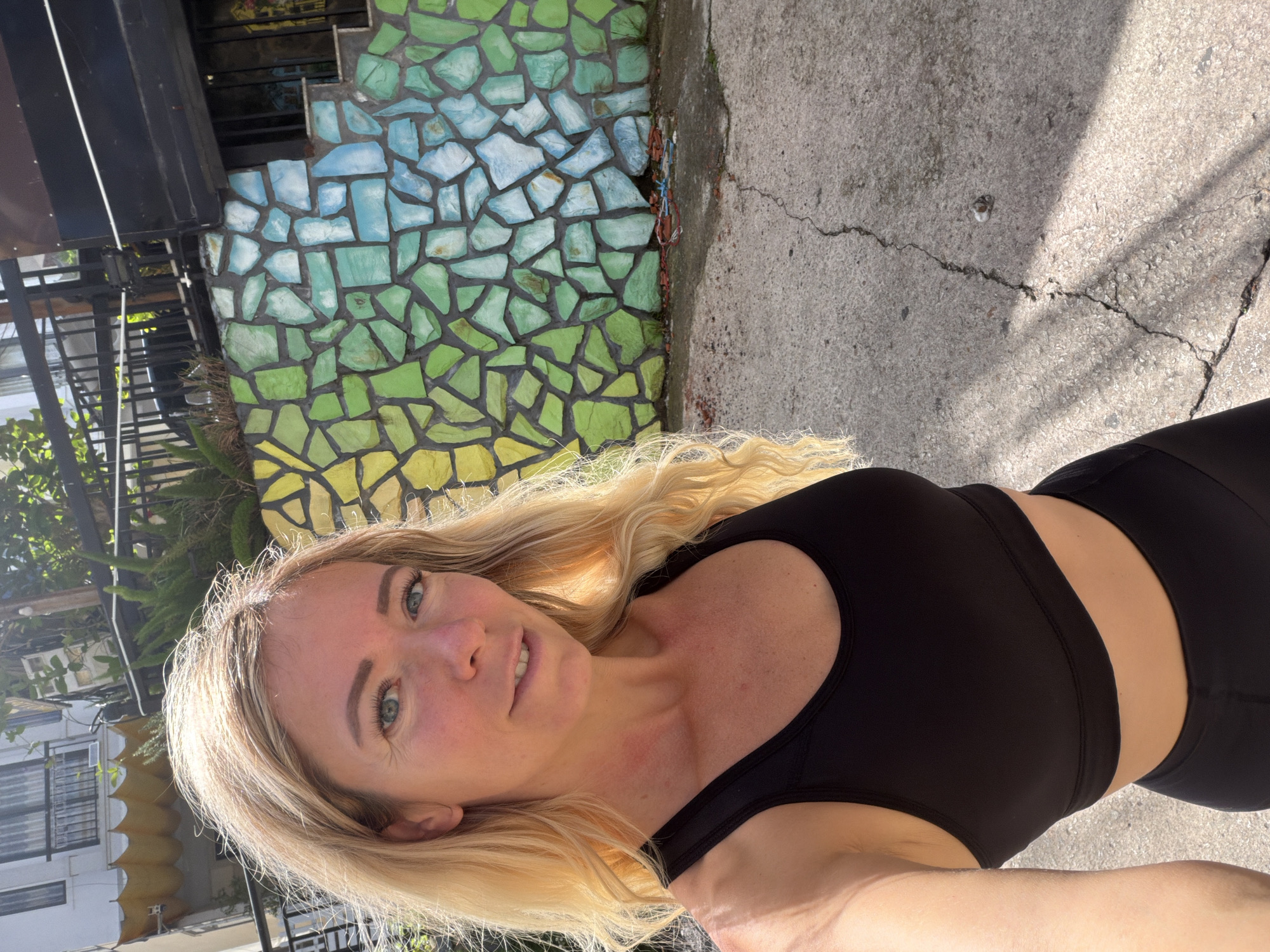 A blonde woman with blue eyes smiles at the camera in a close-up outdoor shot. She is wearing a black sports bra and black leggings. Behind her, a colorful mosaic wall is visible, with green and blue tiles. The ground is made of cracked concrete, and sunlight casts shadows. The background suggests an urban environment with buildings and foliage. The image is a selfie taken during the daytime in Da Lat, Vietnam.