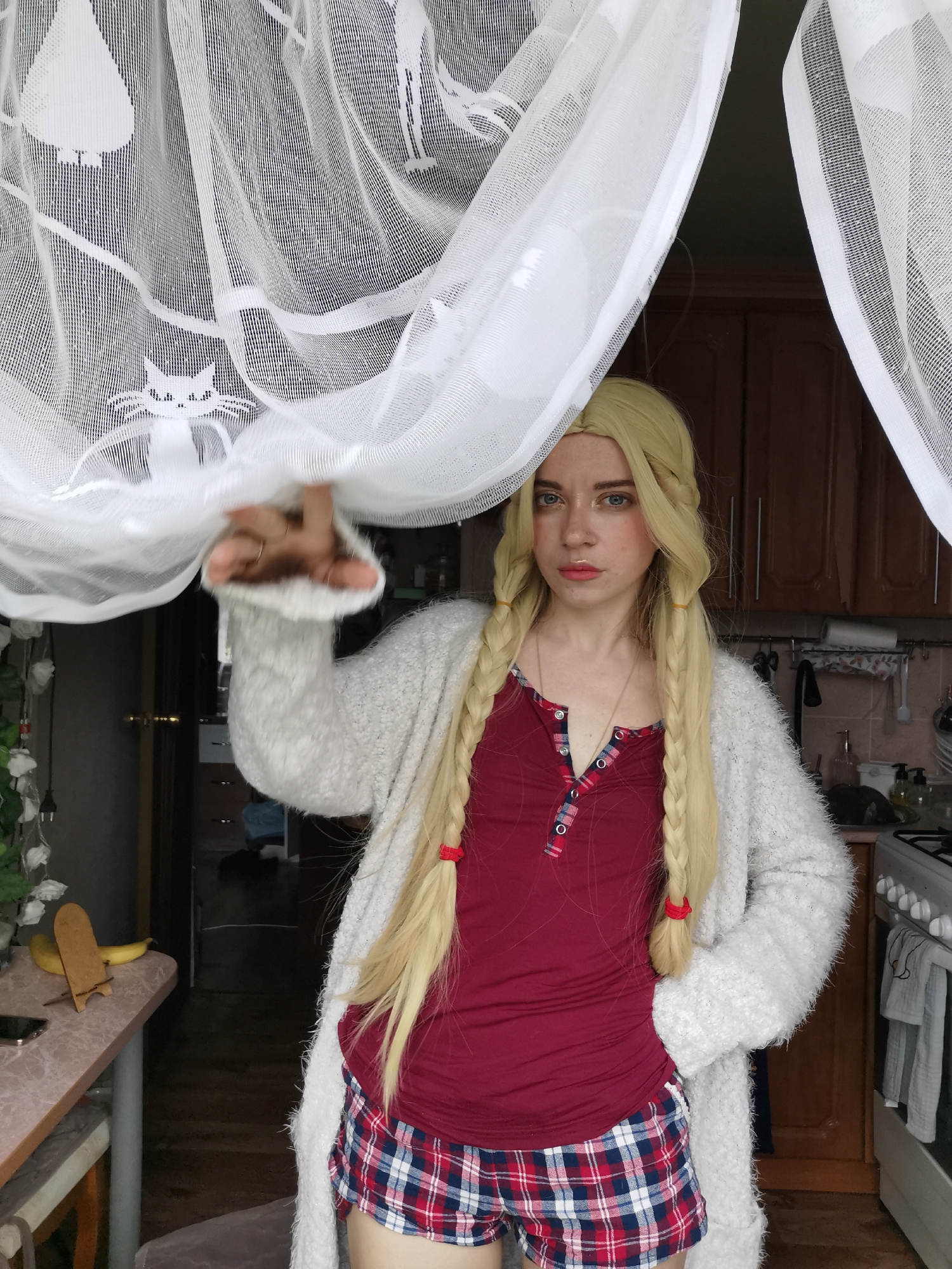 A woman with blonde hair in two braids, wearing a red shirt with buttons and a fluffy grey cardigan over plaid shorts, stands in a kitchen. She is partially obscured by sheer white curtains patterned with silhouettes of cats. The kitchen has wooden cabinets, a stove, and a counter with a banana on it. The overall setting appears to be an indoor domestic environment.