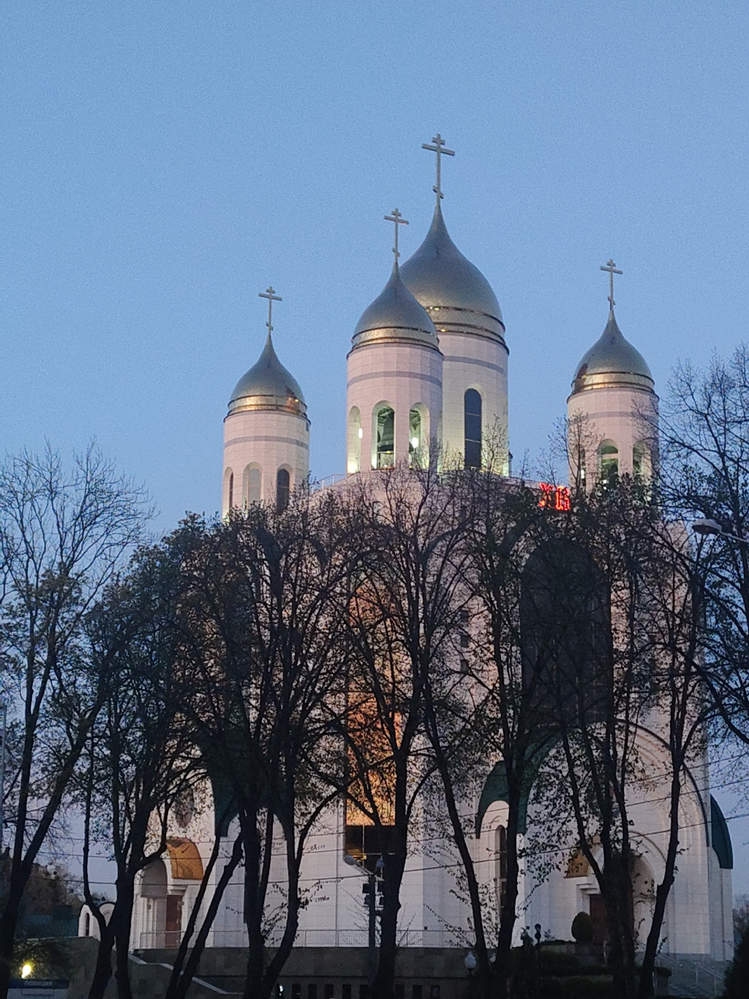 The image features a majestic Eastern Orthodox church at twilight or early evening, characterized by its white facade and multiple golden, onion-shaped domes, each topped with an Orthodox cross. The sky above is a clear, deep blue.
In the foreground, a dense network of bare deciduous tree branches partially obscures the lower and mid-sections of the church, indicating a late autumn or winter setting. Warm light emanates from within some of the church's arched windows, and parts of the building, especially around the domes and bell towers, are illuminated.
A distinct red illuminated sign is visible on the upper section of the church's main structure. The text appears to be in Cyrillic letters, specifically "H" above "ET", possibly forming "HET" if read horizontally. No people are visible, nor is any specific activity taking place, but the illumination suggests the building is active or prepared for services. The scene conveys a sense of grandeur and peaceful solemnity.