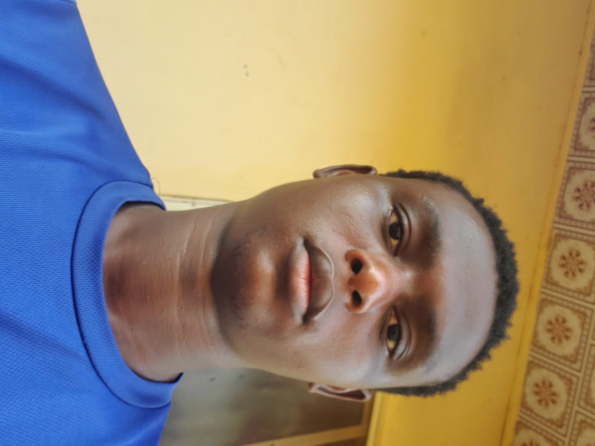 A young black male, wearing a bright blue t-shirt, poses for a selfie. He has dark, short, curly hair and brown eyes. The background is a yellow wall with a patterned border at the top. The lighting is bright, casting a subtle shadow on his face. The photo appears to be taken indoors or in a covered outdoor area. The image is a close-up, focusing on the man's face and upper body.
