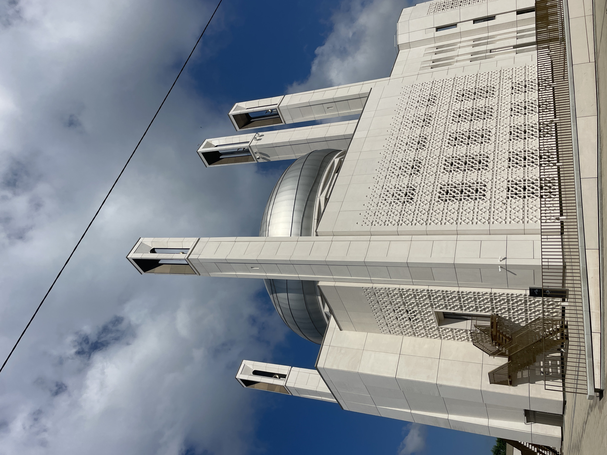 An exterior, low-angle, medium shot captures the modern facade of the Nur Mosque in Almetyevsk, Russia. The mosque features a large, silver dome and four tall, slender towers with open, rectangular tops. The exterior walls are clad in light-colored stone tiles, with intricate geometric patterns carved into sections of the facade. A metallic staircase, painted gold, ascends to an upper level on the left side of the building. A dark metal fence encloses the foreground area. The sky above is a vibrant blue with scattered white and grey clouds, and a black wire runs diagonally across the upper portion of the frame. The scene is devoid of any people.