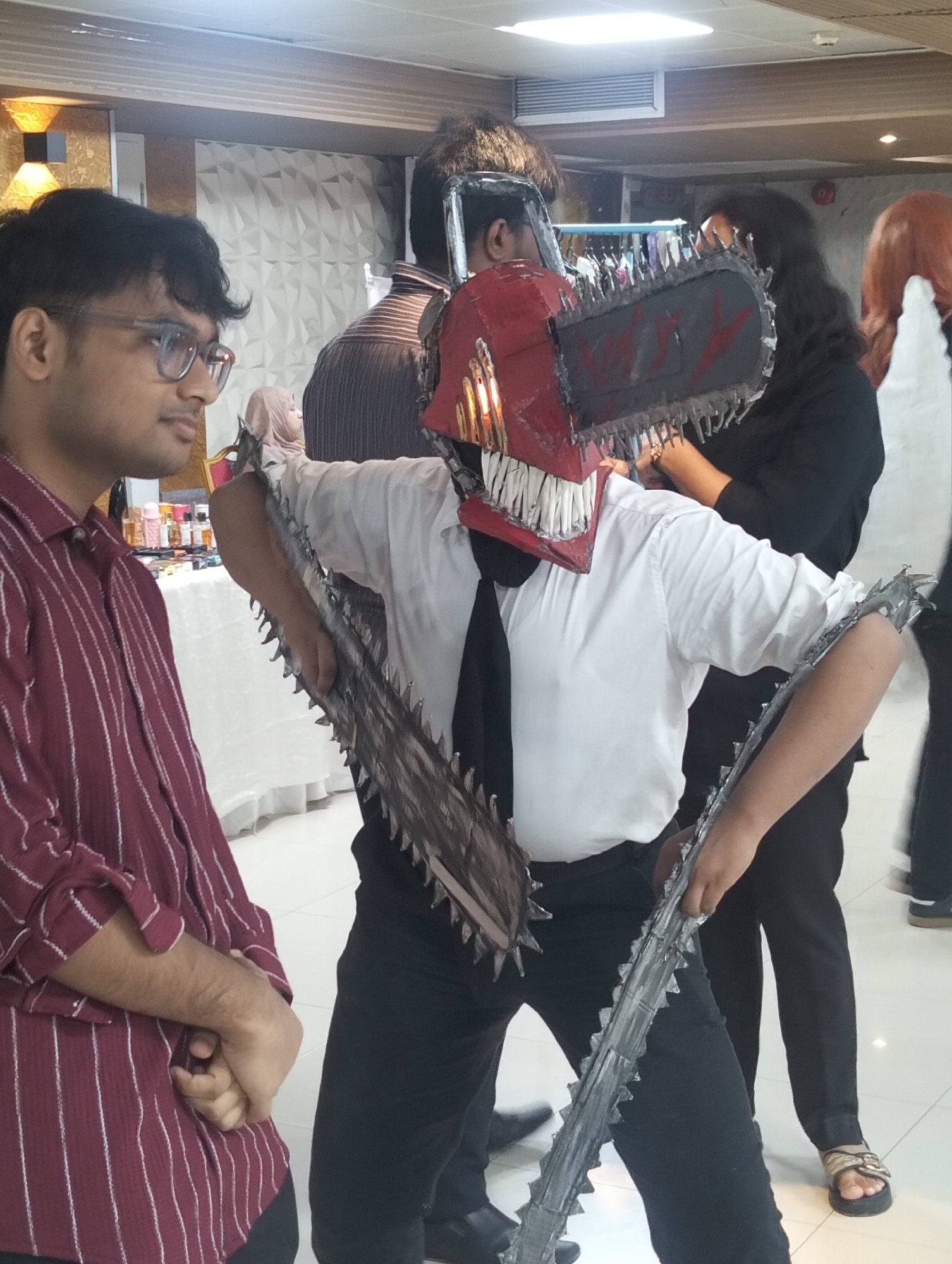 In a bright indoor setting, likely a convention or event space, two individuals are prominently featured. On the left, a young man wearing glasses and a red and white striped button-down shirt stands looking to the right. In the center, a person in a white shirt and black tie is dressed in a striking costume. This costume includes a red chainsaw-like mask with sharp teeth and two large, serrated chainsaw blades extending from their arms and shoulders. The background reveals a white table laden with various items, possibly merchandise or displays, and other individuals, some with obscured faces. The flooring is light-colored tile. The overall atmosphere suggests a public gathering with a focus on cosplay or themed entertainment.