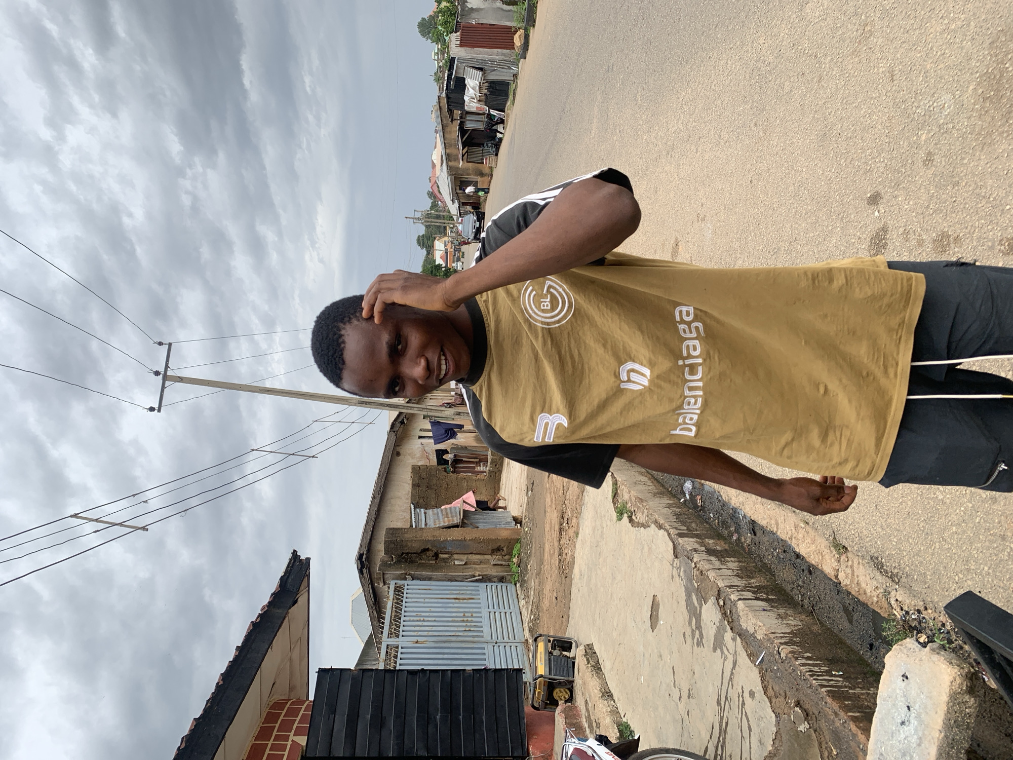 A young Black male stands on an asphalt road in a residential area of Jos, Nigeria. He is wearing a mustard yellow t-shirt with black sleeves and black cargo shorts. His right hand is raised to his temple, and he is looking at the camera with a slight smile. The t-shirt has a graphic logo on the chest, which appears to be a circle with the letters "BL" inside, and the word "Balenciaga" is visible near the bottom hem.
Behind him, the road is lined with modest buildings constructed from various materials, including corrugated metal, concrete blocks, and some with brick facades. Some buildings appear to be shops or workshops, with open fronts and goods displayed. Clothes are hanging on a line outside one of the structures. A blue metal gate is visible to the left of the man, in front of a brick building with a black-tiled roof. A yellow generator is placed on a concrete walkway next to the gate. Overhead, electrical wires stretch across the frame, supported by wooden poles. The sky is overcast with a dull, grey cloud cover. The ground on the side of the road consists of a concrete walkway and a shallow drainage ditch filled with debris. A motorcycle is partially visible in the lower left corner of the image.