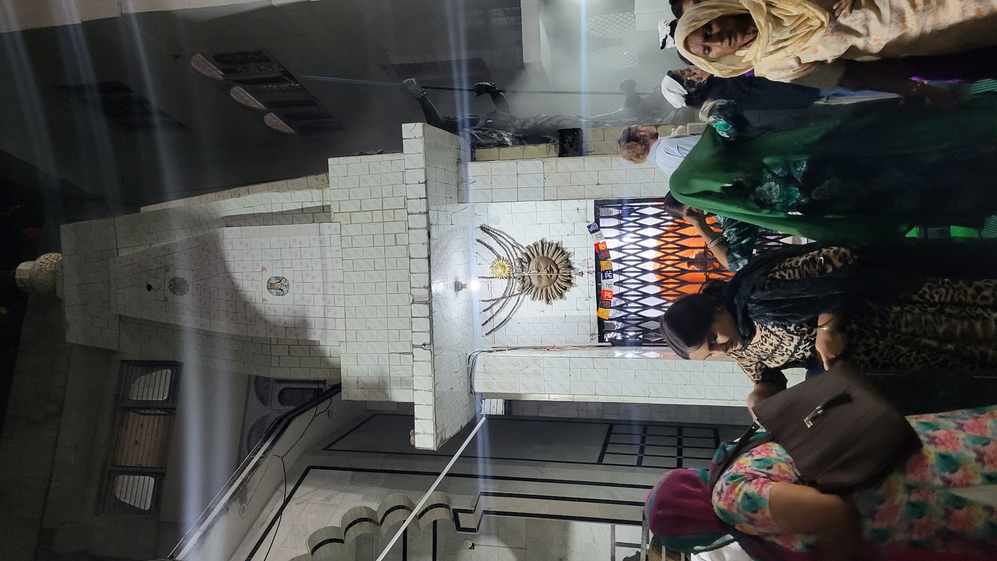 This image, captured in Kasol, India, portrays a gathering of people within what appears to be a temple or spiritual center. The setting is characterized by light-colored tiled walls, some featuring decorative square tiles with subtle motifs. A central feature is a prominent sun-like sculpture mounted on the wall, adorned with radiating chains or rays, and illuminated by a light fixture beneath it. To its right, a patterned metal gate or screen is visible, decorated with small flags bearing text in a Devanagari-like script, possibly Hindi or Nepali, with discernible symbols such as 'Om'.
Several individuals are present, mainly women dressed in traditional South Asian clothing. In the foreground, a woman in a leopard-print dress and black shawl is seen looking downwards, holding a dark brown bag. Partially visible behind her is another woman in a floral-patterned top with a pink hood. Further back, a woman in a green dress or sari stands alongside a woman wearing a light-colored headscarf, who gazes calmly towards the viewer. A man with a beard and a light shirt is also present in the background. On an upper ledge to the left, a dark, abstract human-like figure or sculpture is lying.
The activity suggests people are observing, waiting, or participating in a quiet event or visit at this cultural or religious site. The lighting indicates an indoor setting or evening time under artificial illumination. The overall atmosphere appears tranquil and respectful.