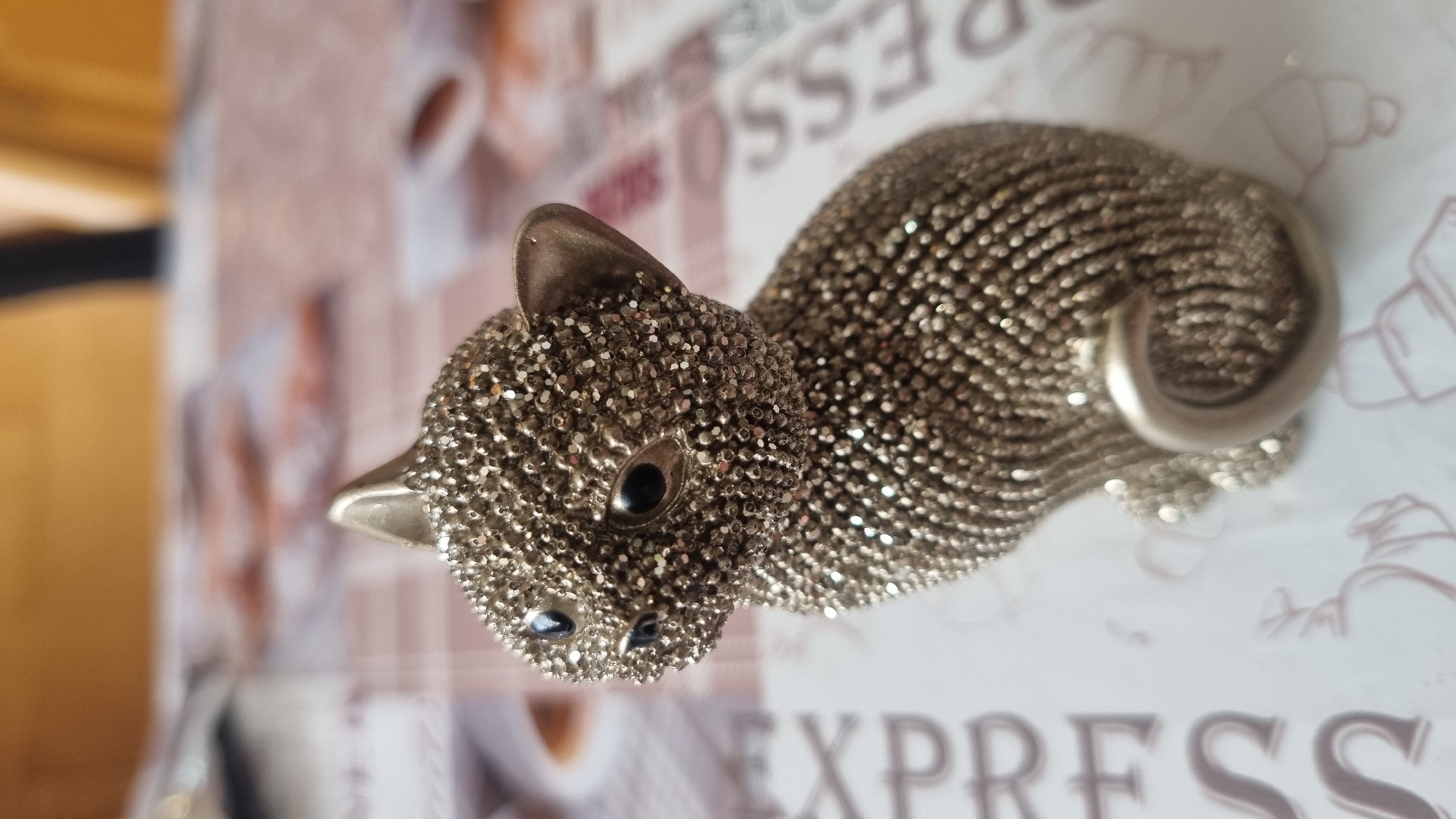A small, silver, bejeweled cat figurine is centrally placed on a surface covered with a patterned tablecloth. The tablecloth has a light pinkish hue and features faint illustrations of coffee cups and bags. To the left of the figurine, the word "EXPRESS" is visible in large, stylized lettering. To the right, parts of the word "ESSES" can be seen in similar lettering. The background is out of focus, showing a warm, golden-brown color, possibly a wooden surface or wall. The lighting suggests an indoor setting, likely a table in a room. There are no people present in the image. The image itself is a photograph taken from a slightly elevated angle, focusing on the figurine. The origin of the image, specifically within the city of Smila, Ukraine, cannot be determined from the visual information provided.