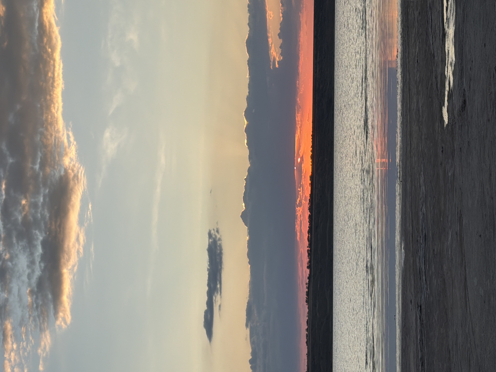 The image depicts a tranquil sunset over the Kuyalnik Estuary in Odesa, Ukraine. The sky is a canvas of soft blues, oranges, and pinks, with clouds illuminated by the setting sun. A strip of land, possibly the estuary's bank, is visible in the middle ground, silhouetted against the vibrant sky. The water of the estuary reflects the colors of the sunset, creating a mirror-like effect. The foreground shows a sandy, somewhat barren shoreline, with evidence of shallow water puddles. No people or specific structures are visible, suggesting a natural landscape setting.
