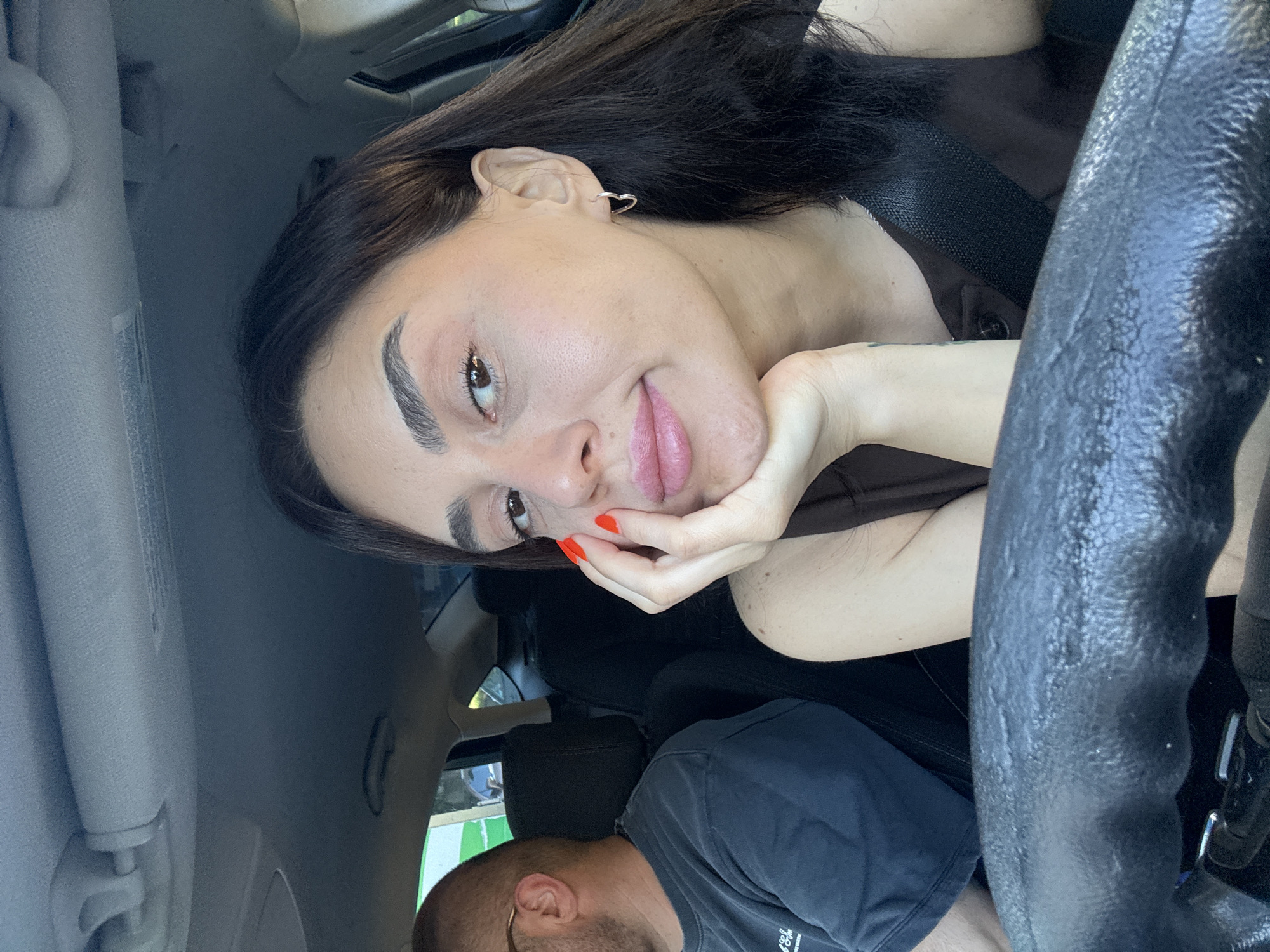 A woman with dark hair, wearing a dark brown top and a seatbelt, rests her chin on her hand, her painted red nails visible. She is in the passenger seat of a car, with the steering wheel in the foreground. A man with glasses and a dark t-shirt is visible in the driver's seat on the left. The background shows a street scene through the car window. The image is a candid, natural light photograph.