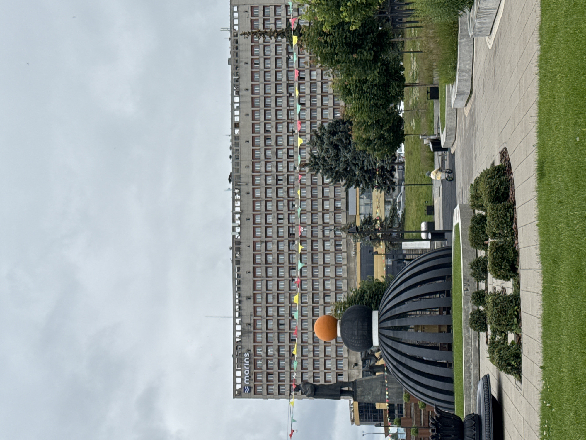 The image depicts a scene in Nizhny Novgorod, Russia, dominated by a large, Brutalist-style hotel, likely the "Marins Park Hotel" indicated by signage. In the foreground, there's a dark, semi-spherical monument with an orange sphere on top, surrounded by manicured greenery and paved walkways. A statue of a man stands to the left of the monument. Festive banners with triangular flags are strung across the scene, suggesting a celebratory occasion. A child on a small bicycle is visible in the background, riding on a pathway. The sky is overcast, contributing to a muted color palette. The overall environment suggests a public square or park area within the city.