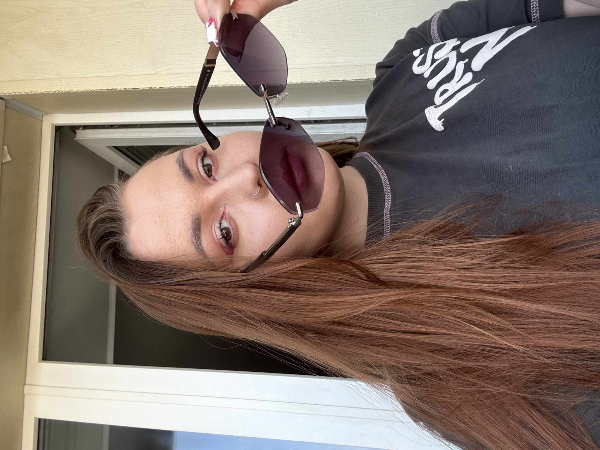A woman is holding a pair of rimless sunglasses with dark lenses. She has long brown hair and a nose ring. She is wearing a dark gray t-shirt with white lettering. The background appears to be a window frame and an exterior wall. There is no information to suggest the location is Izhevsk, Russia.